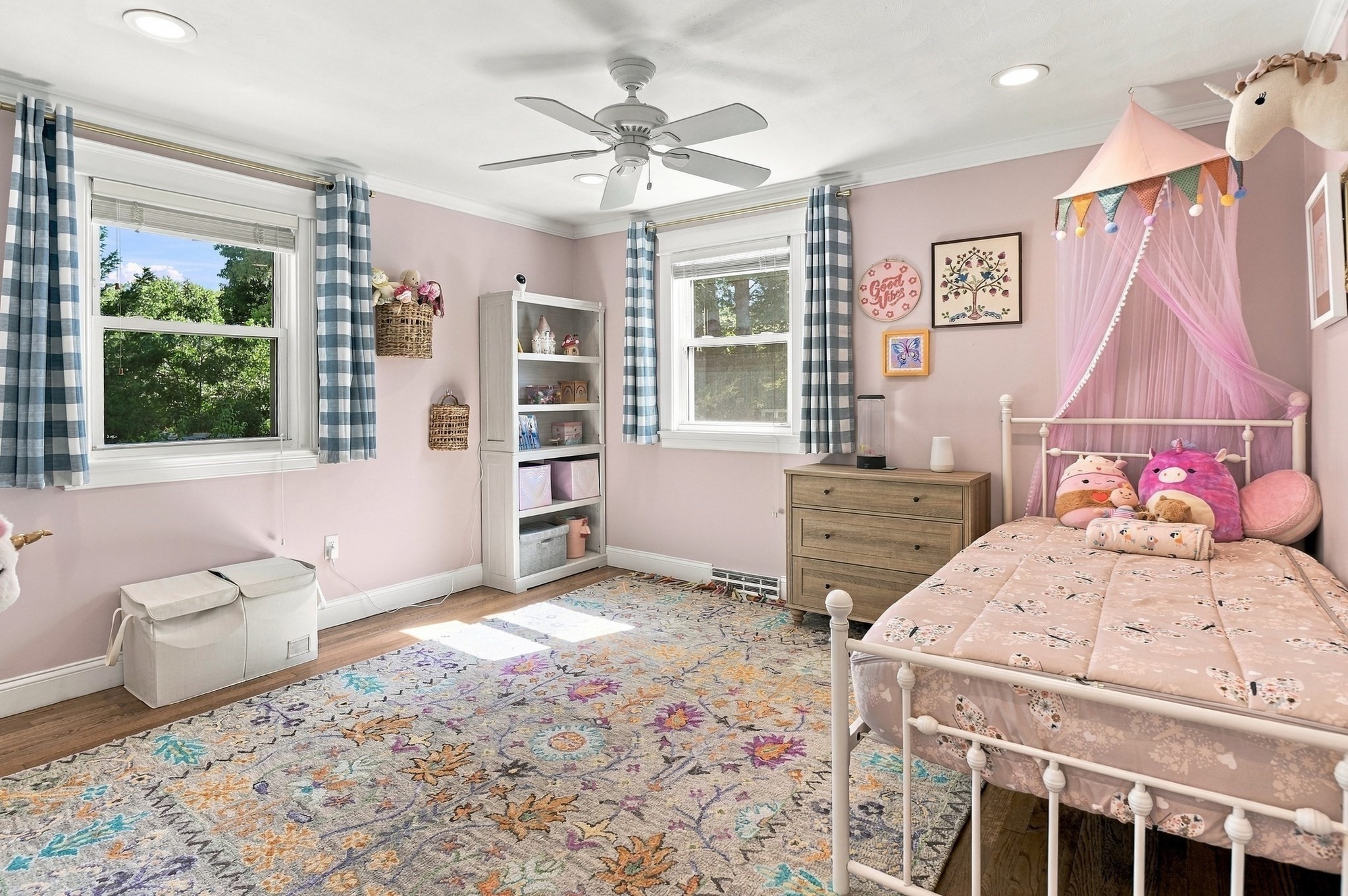 Pink child's bedroom with a canopy bed, plush toys, checked curtains, and a floral rug.