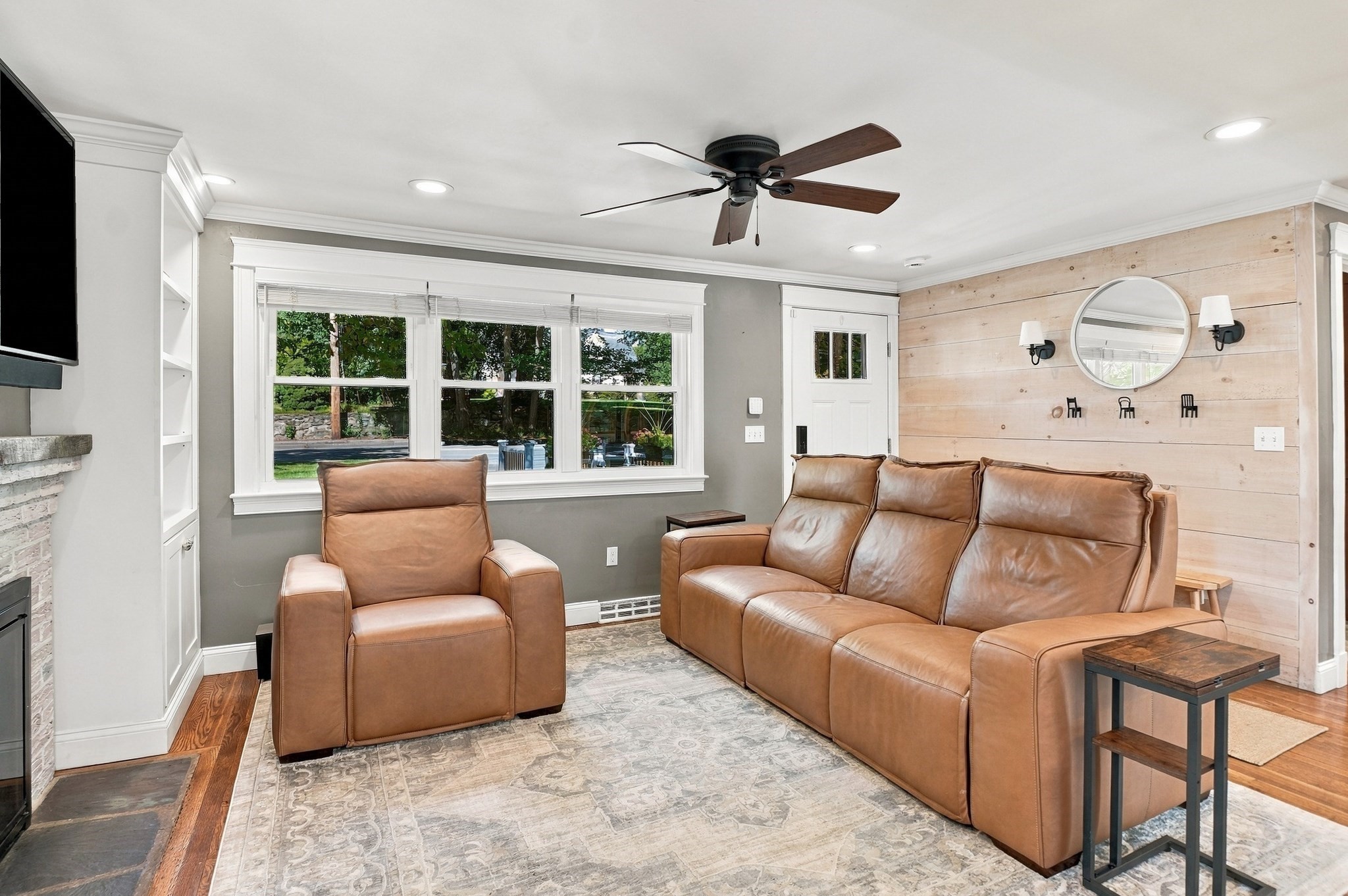 Living room with tan leather seating, a pale wood accent wall, large windows, and a stone fireplace.