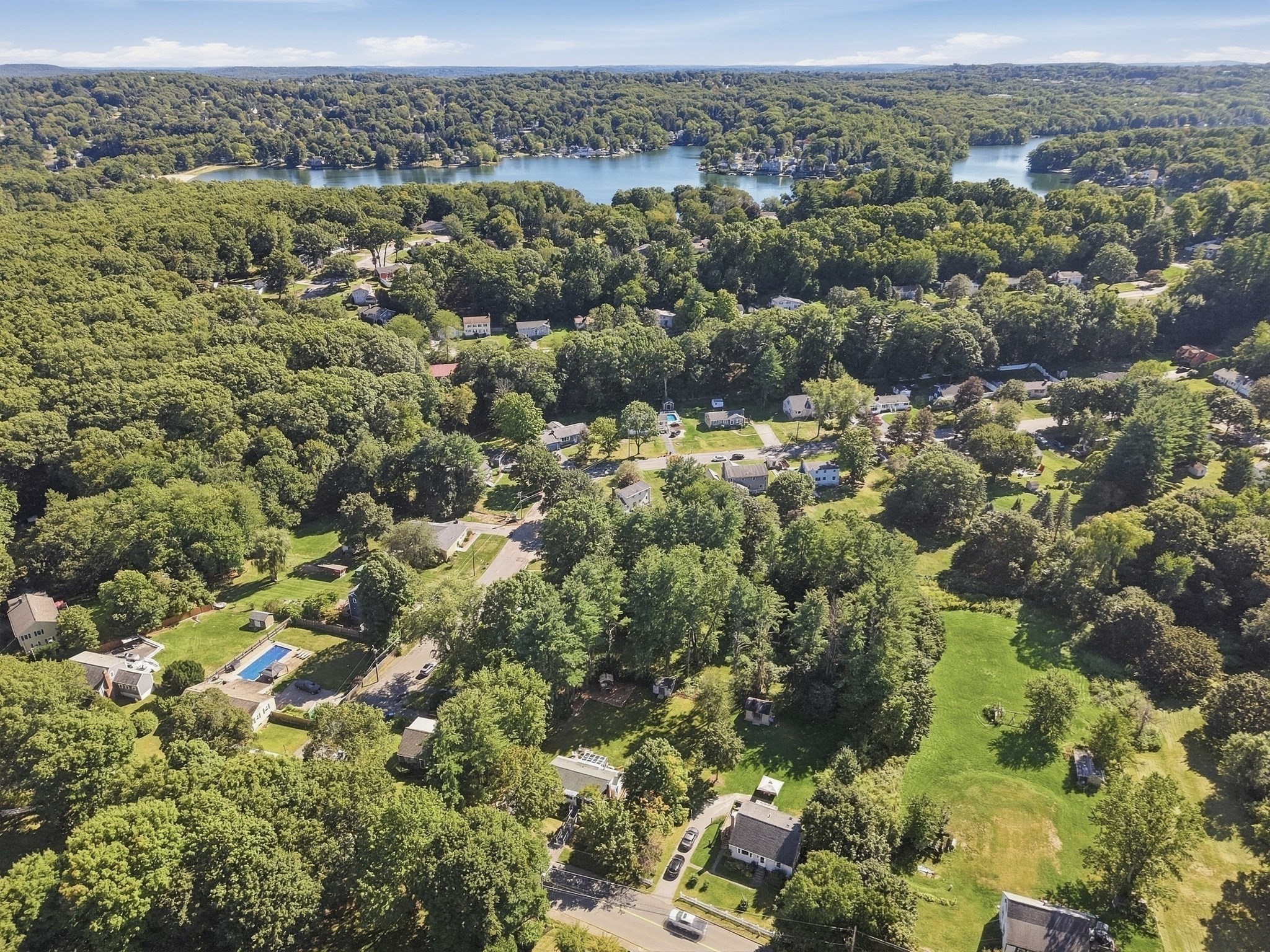 Aerial view of a wooded neighborhood with a lake in the distance and a backyard pool.