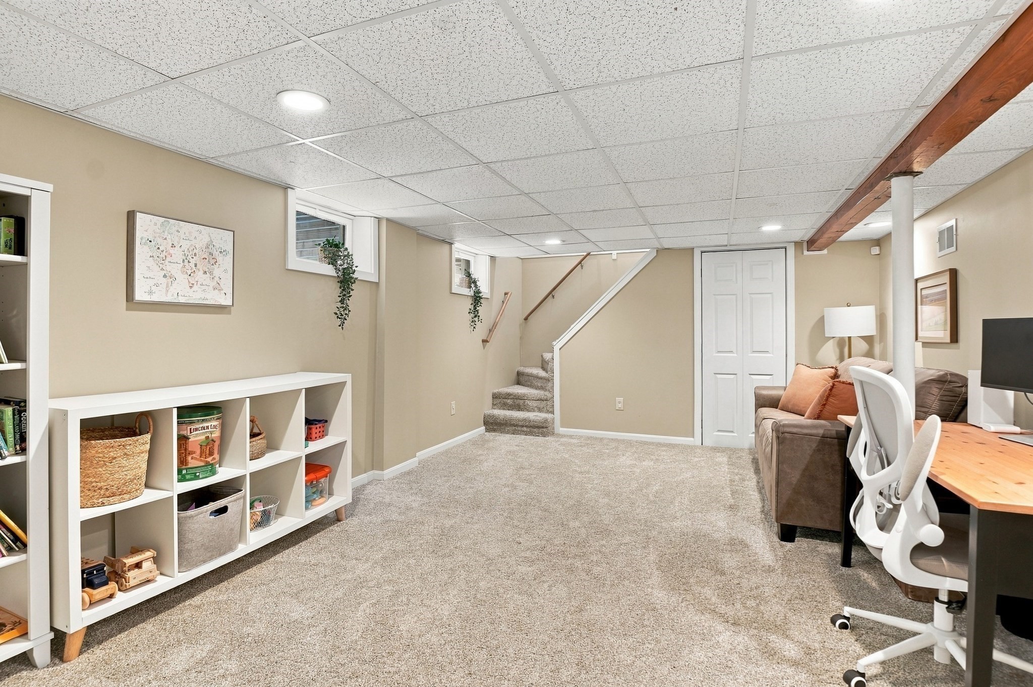 Finished basement with carpet, a staircase, white storage shelves, and a sofa beside a desk.