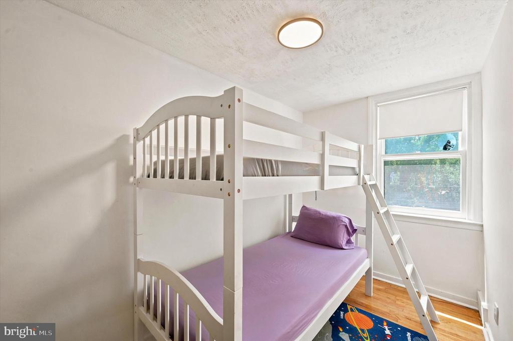 Bedroom with a white bunk bed, purple bedding, and a colorful rug beside a sunlit window.
