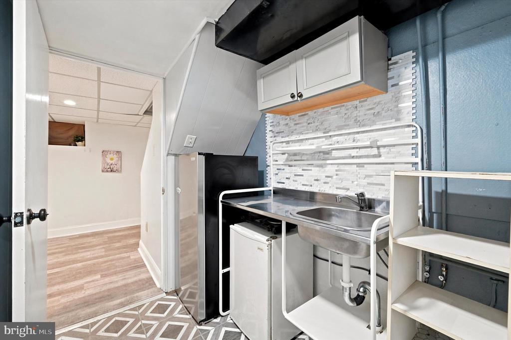Compact kitchenette with a stainless steel sink, mini fridge, white cabinets, and gray-and-white tile backsplash.