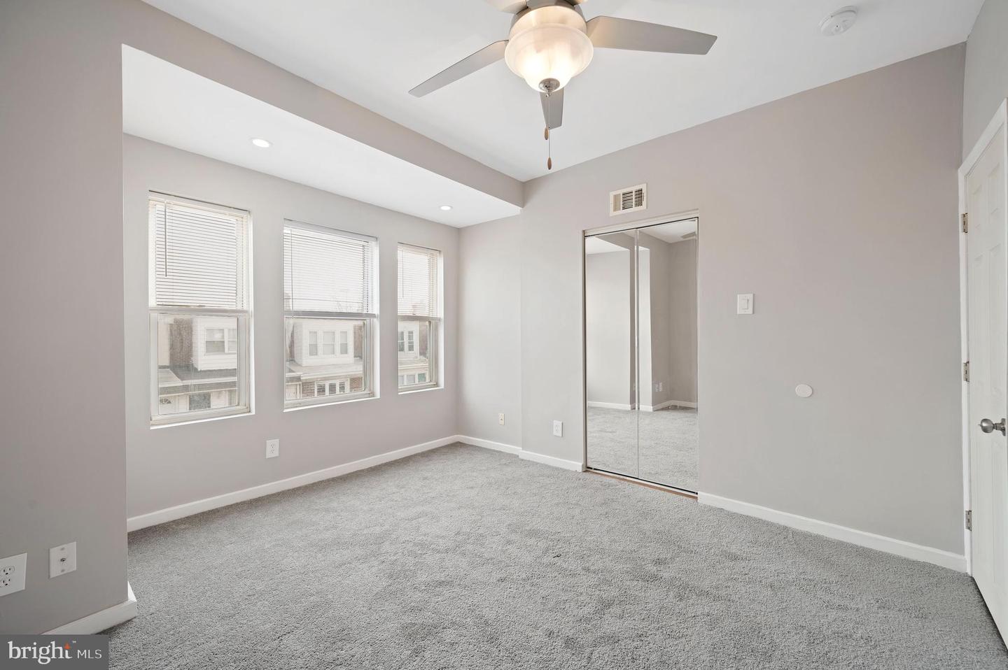 Empty bedroom with gray carpet, three blind-covered windows, mirrored closet doors, and a ceiling fan.