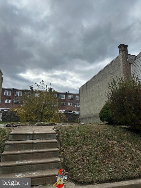 Sloped backyard with concrete steps, patchy grass, shrubs, and a tall side wall.