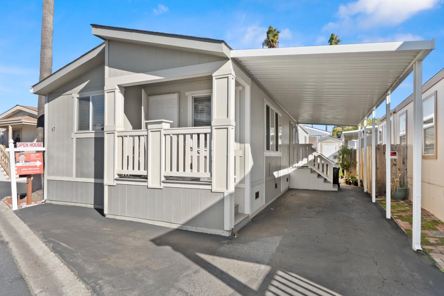 Gray manufactured home with a small porch and long covered driveway beside neighboring homes.