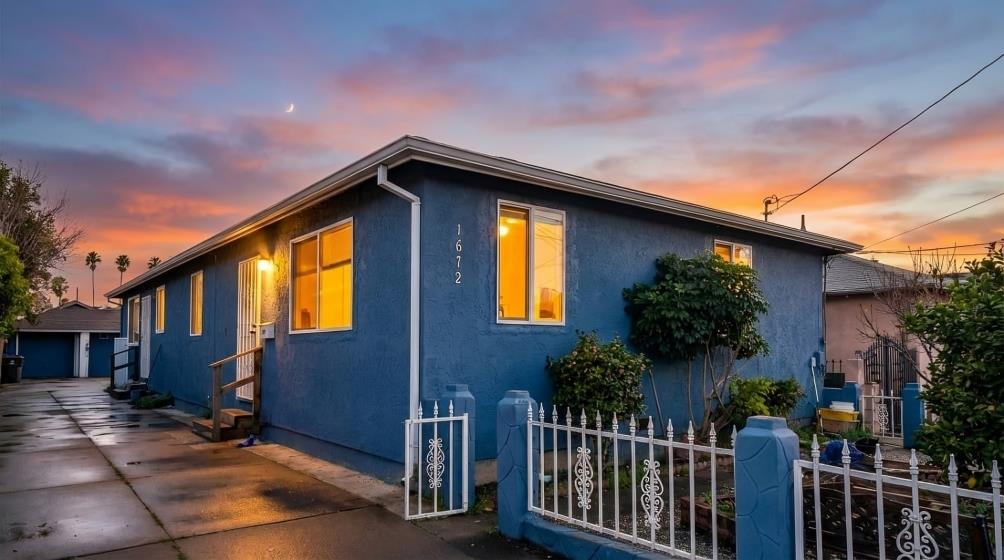 Blue single-story house with a white fence and warmly lit windows at dusk.