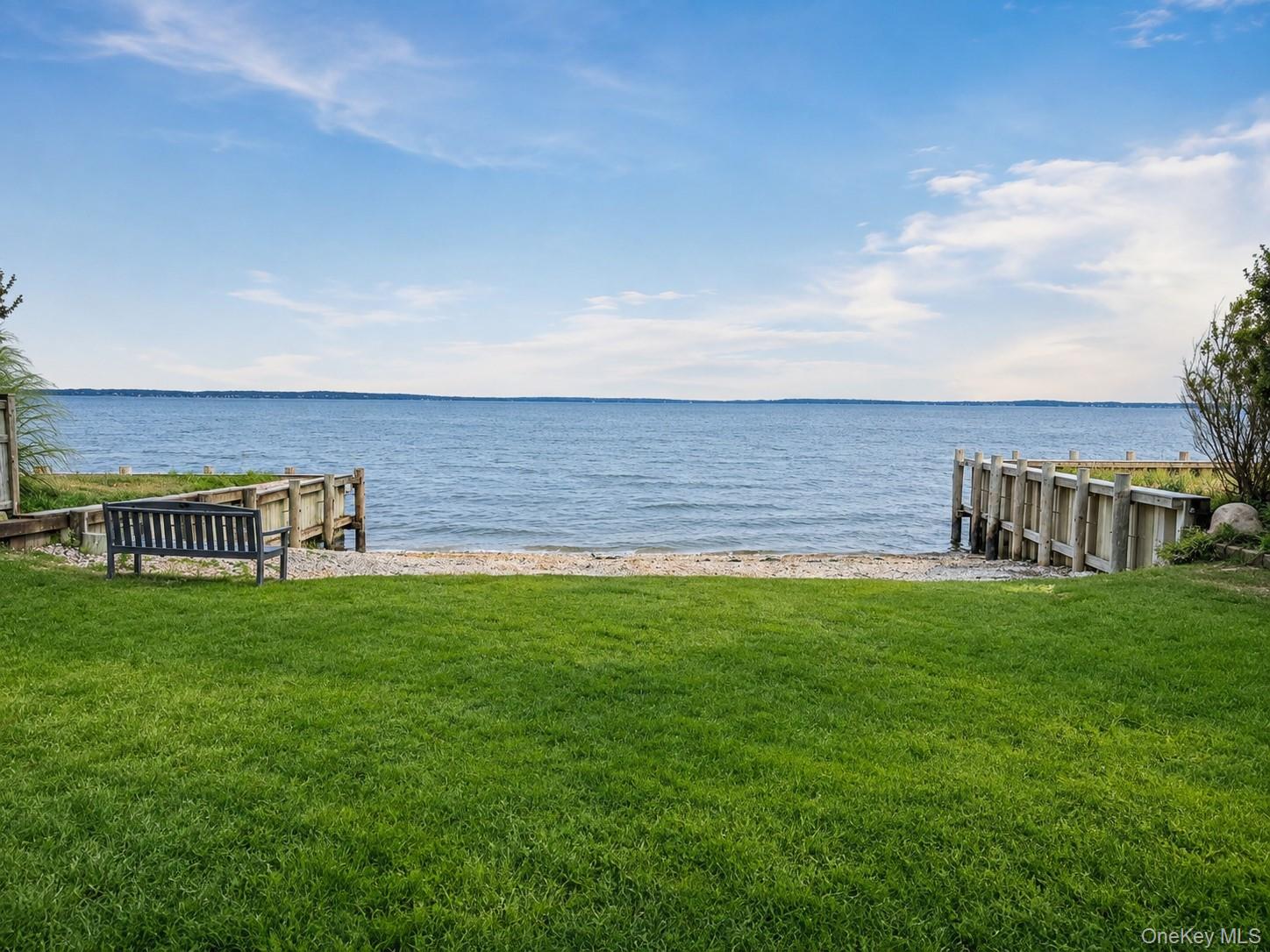 Waterfront lawn leading to a pebbled shore, with a wooden bench and open water beyond.