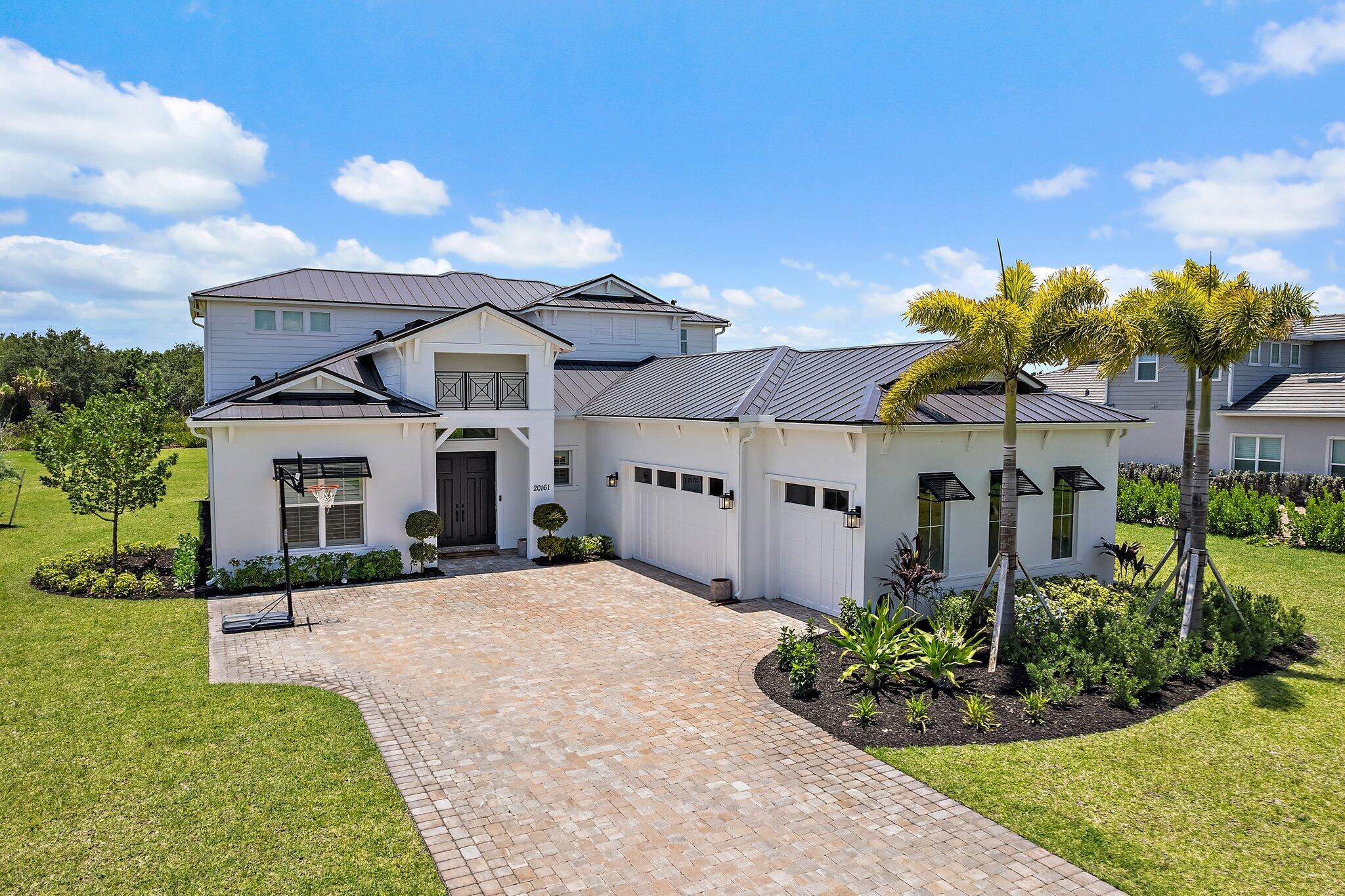 Two-story white home with dark metal roofs, a wide paved driveway, palms, and a basketball hoop.