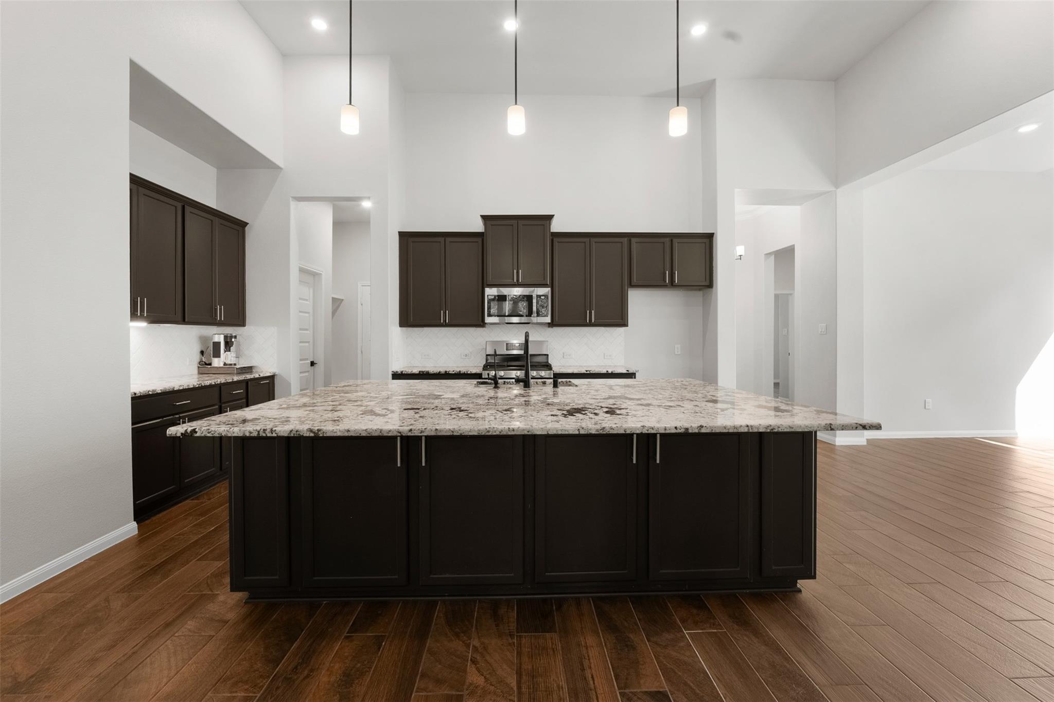Kitchen with a large granite island, dark cabinets, pendant lights, and wood-look flooring.