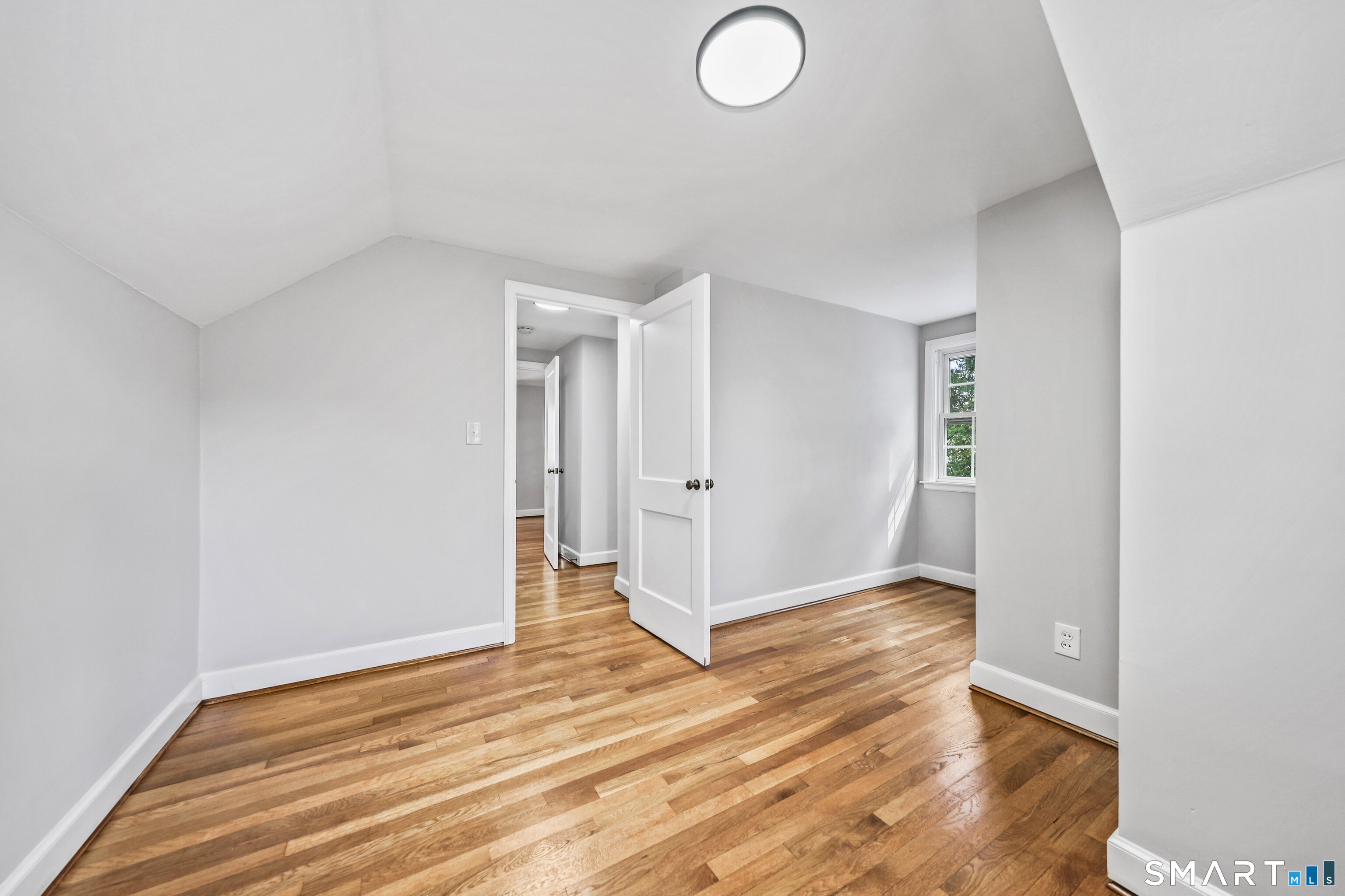 Empty room with hardwood floors, a sloped ceiling, and an open door beside a small window.