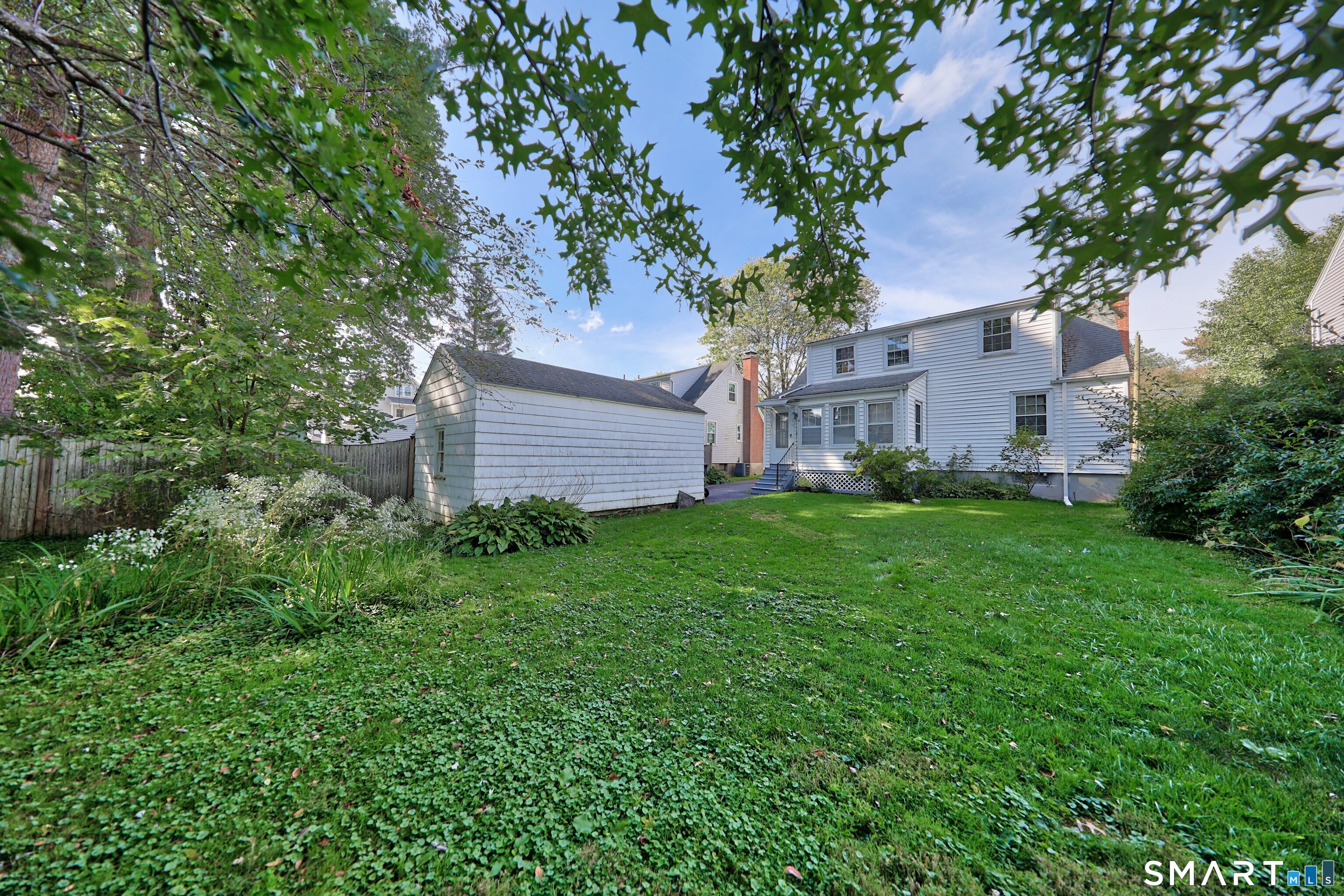 Rear of a white two-story house and detached shed beyond a broad lawn and mature trees.