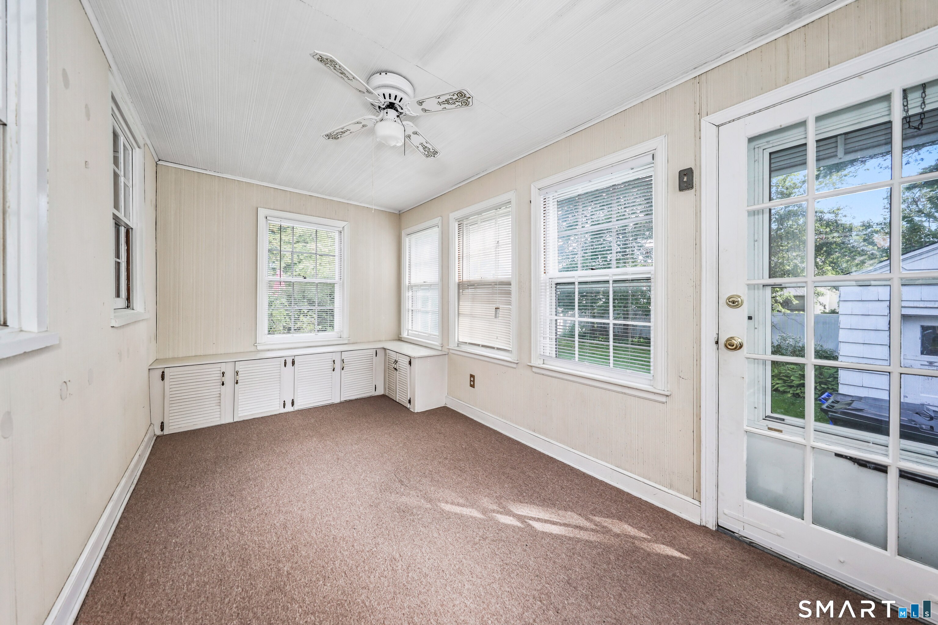 Enclosed porch with brown carpet, built-in louvered cabinets, multiple windows, and a glass-paneled door.