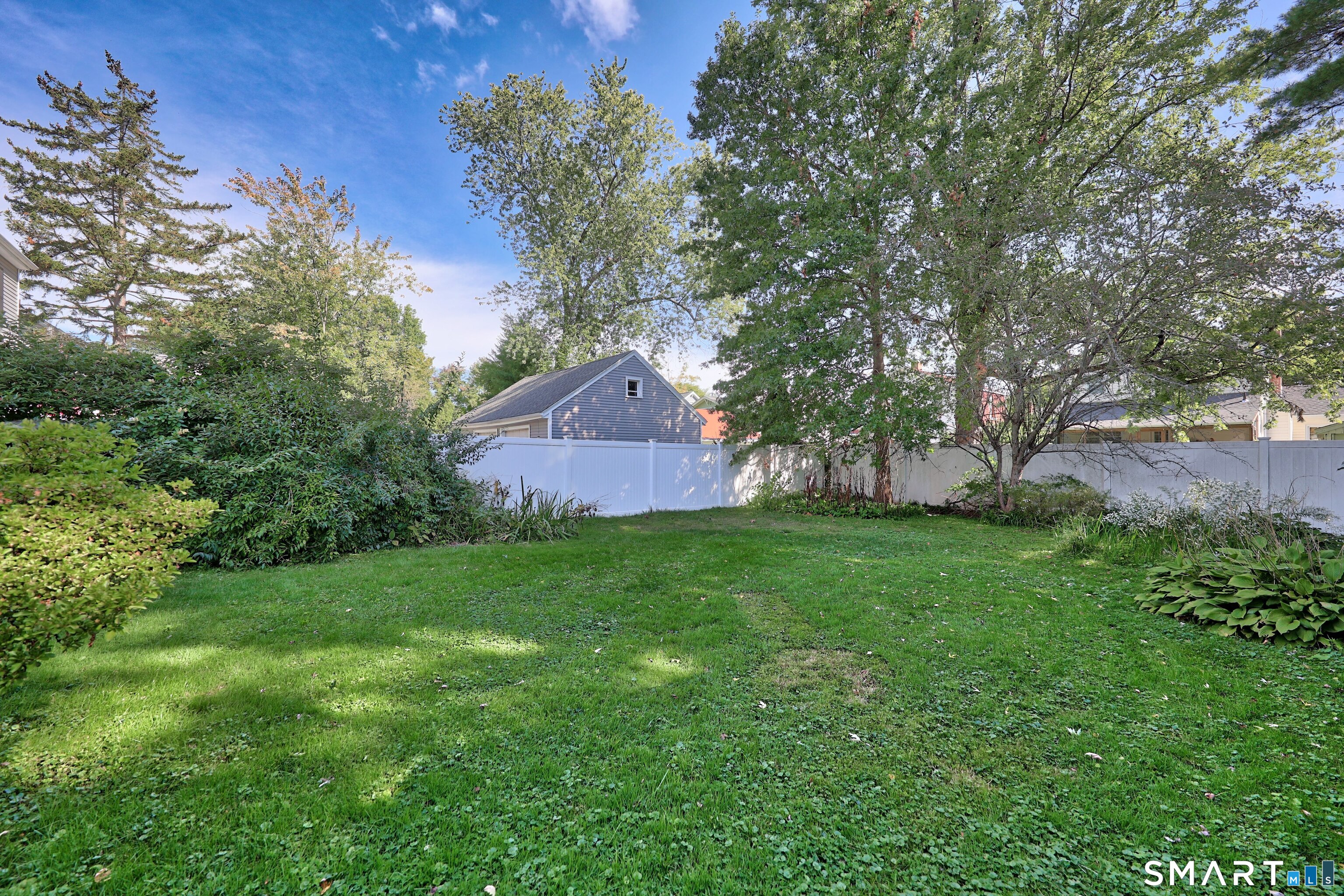 Grassy backyard with mature trees, shrubs, and a white privacy fence.