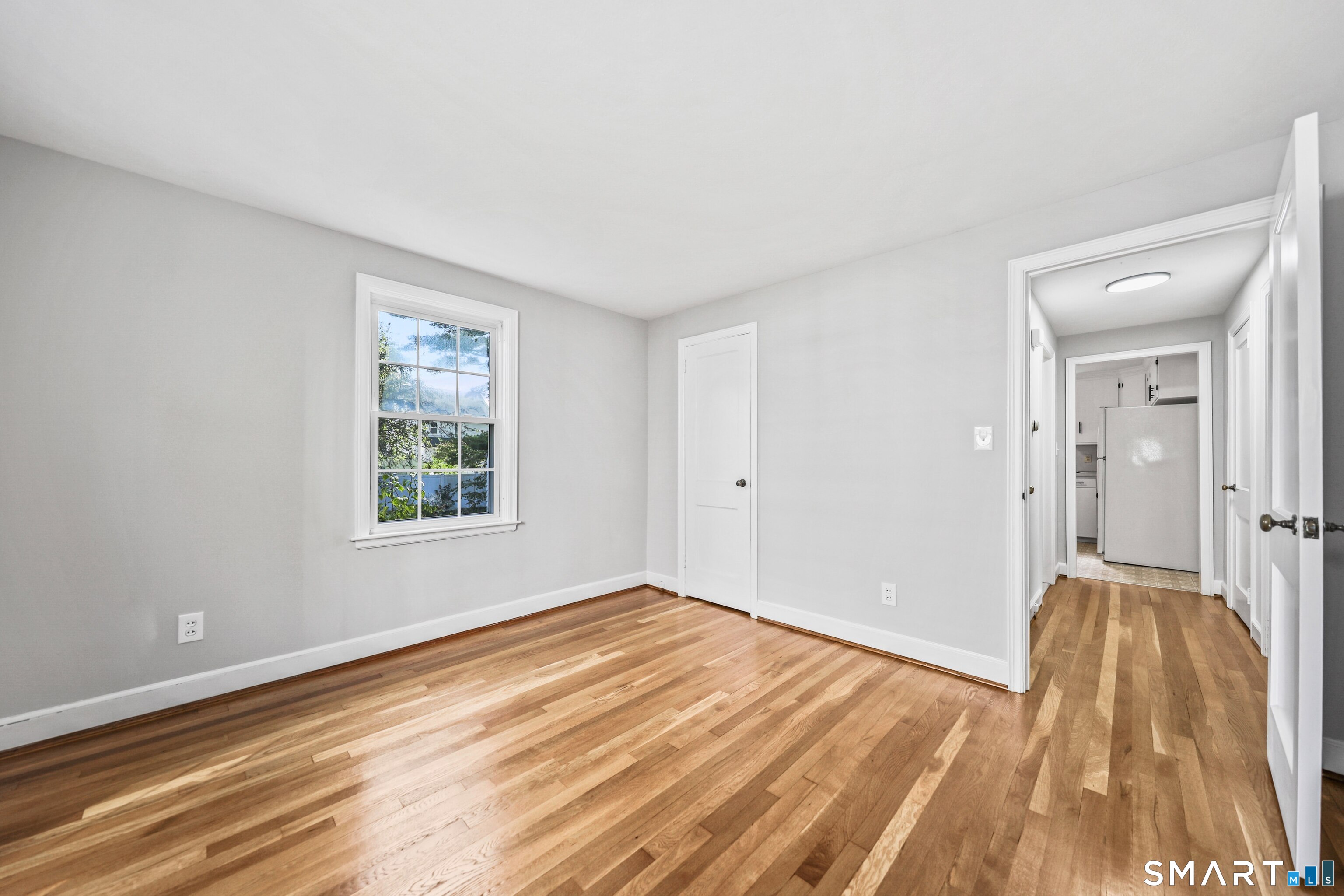 Empty room with polished hardwood floors, a window, and an open doorway to the hall and kitchen.