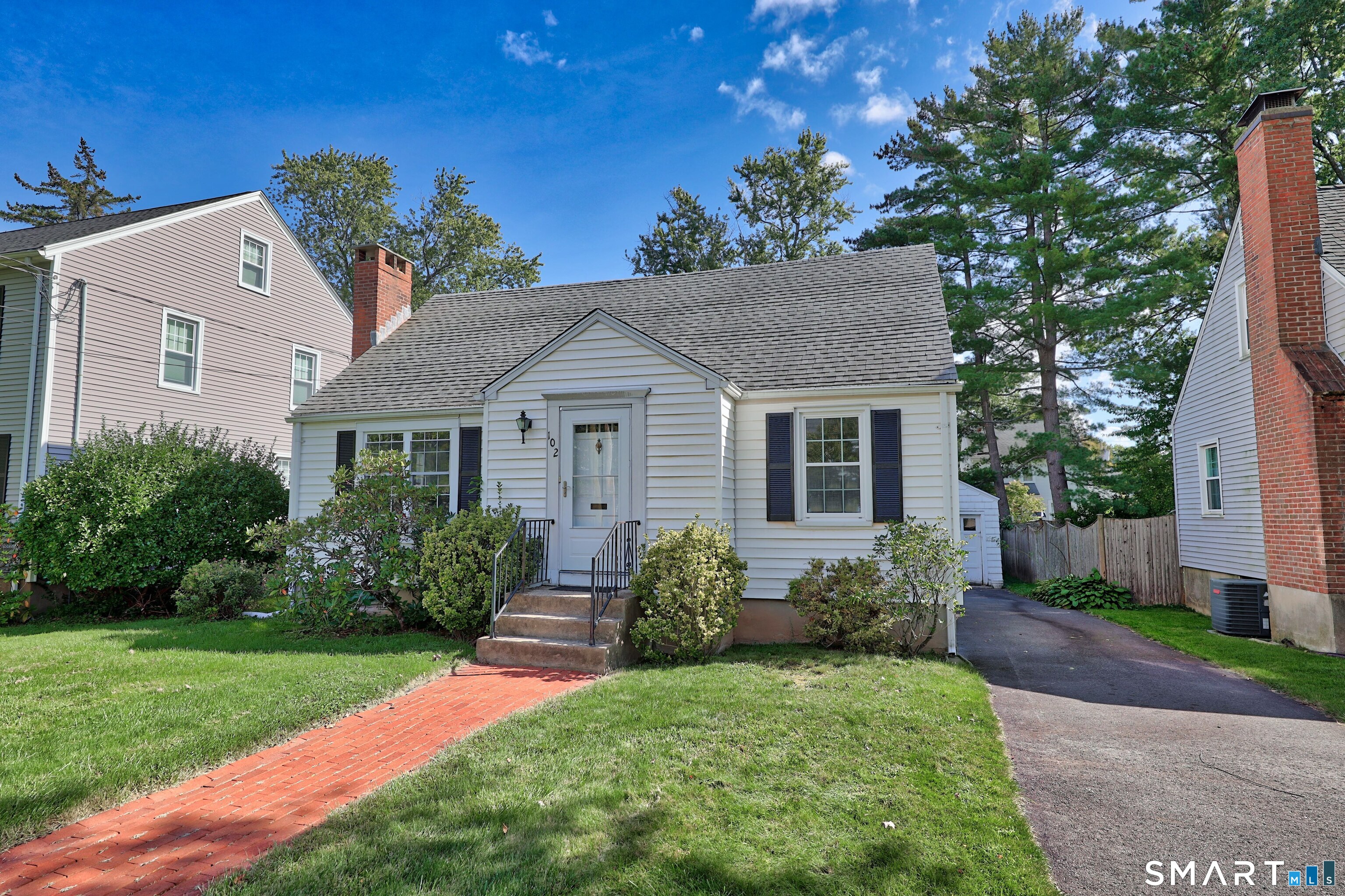 White-sided house with black shutters, brick chimney, front steps, and a brick walkway.