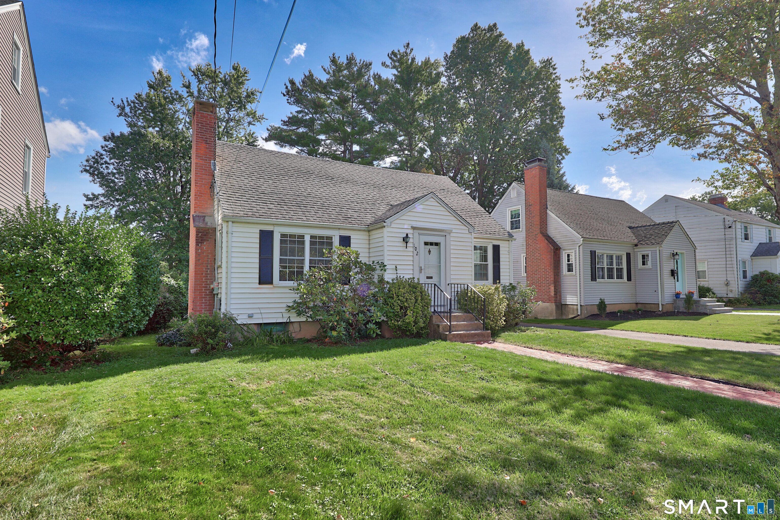 White-sided house with two brick chimneys, front steps, shrubs, and a broad green lawn.