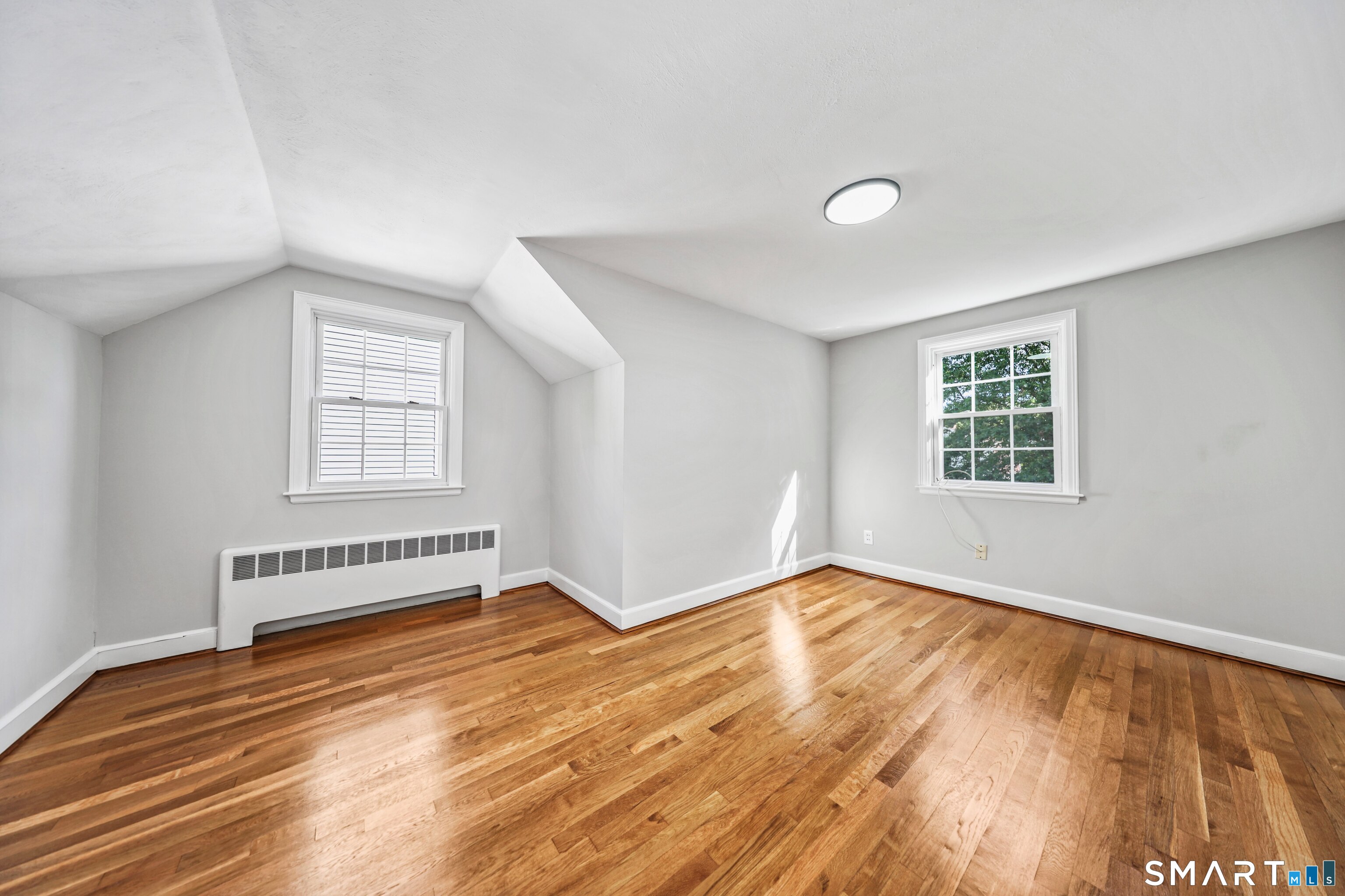 Vacant room with angled ceilings, two windows, a radiator, and polished hardwood floors.