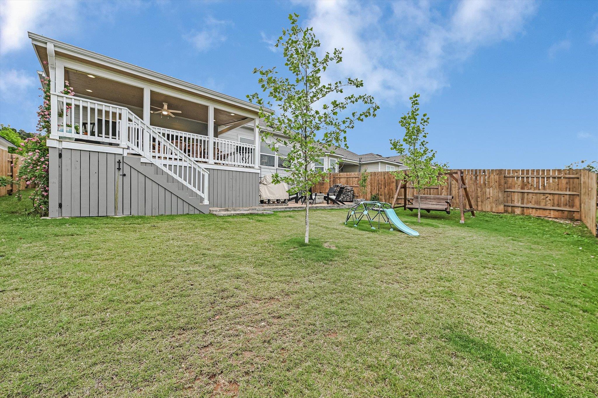 Home’s covered rear deck overlooks a fenced lawn with young trees and a children’s play set.