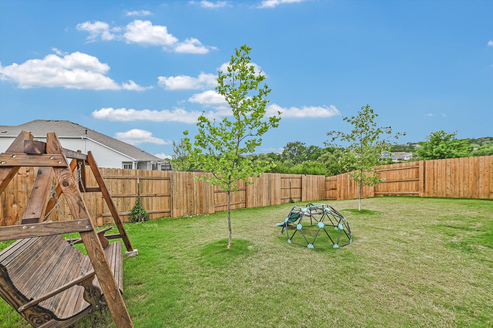 Fenced backyard with a wooden swing set, climbing dome, young trees, and lawn.