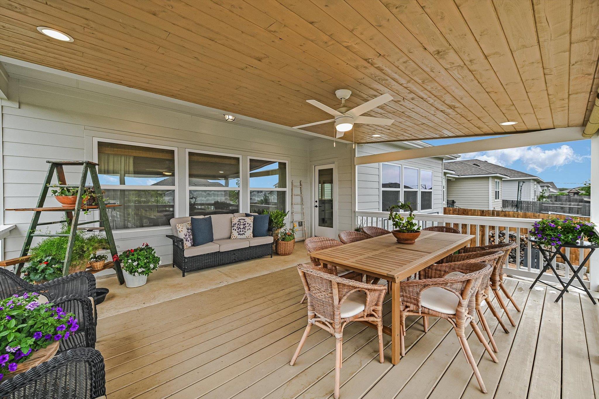 Covered patio with a wood ceiling, wicker dining set, cushioned sofa, and potted flowers.