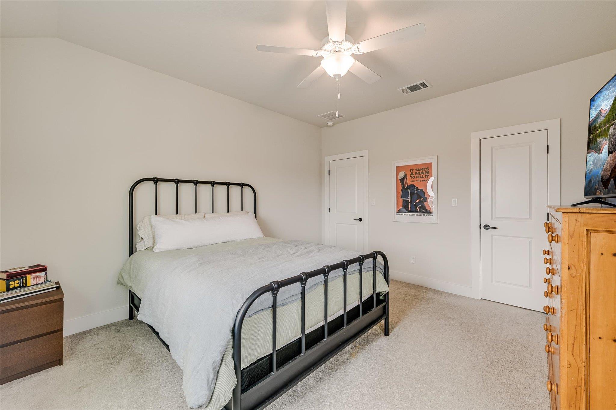 Bedroom with a black metal bed, pale bedding, beige carpet, and ceiling fan.