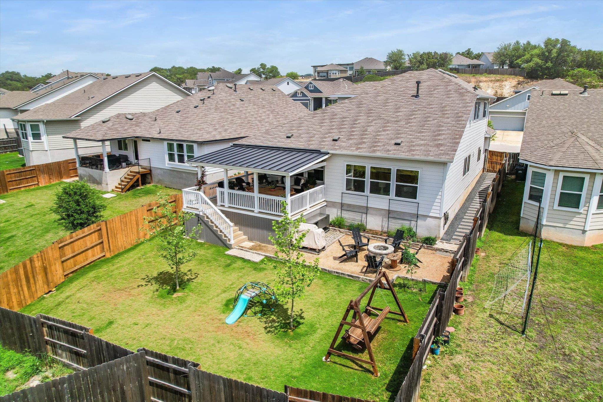 Backyard with a covered deck, fenced lawn, play equipment, and a gravel patio with a fire pit.