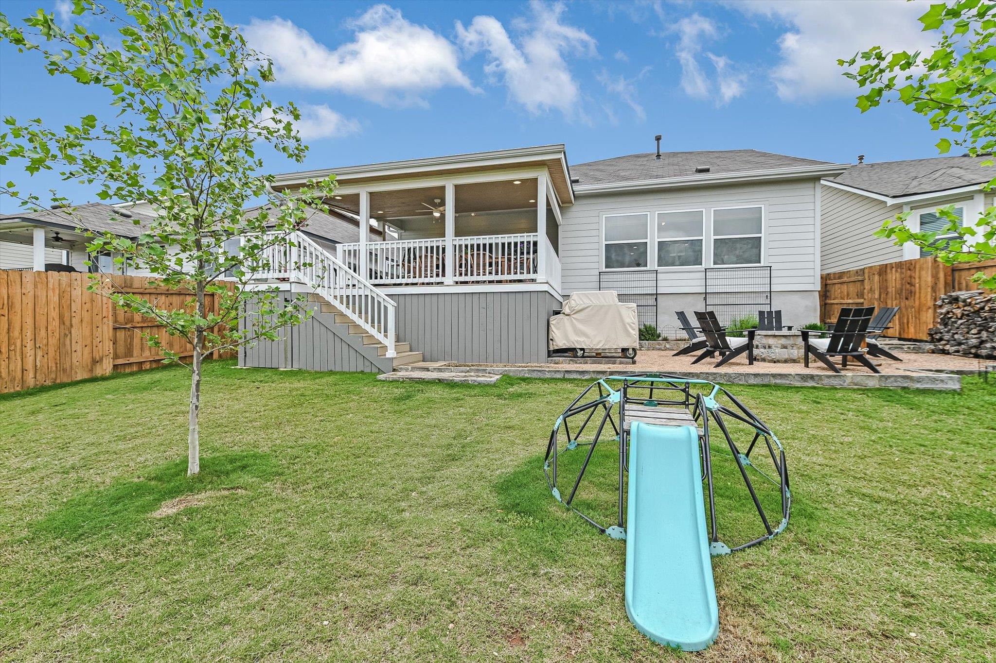 Backyard with a covered raised deck, patio seating, fenced lawn, and blue slide.