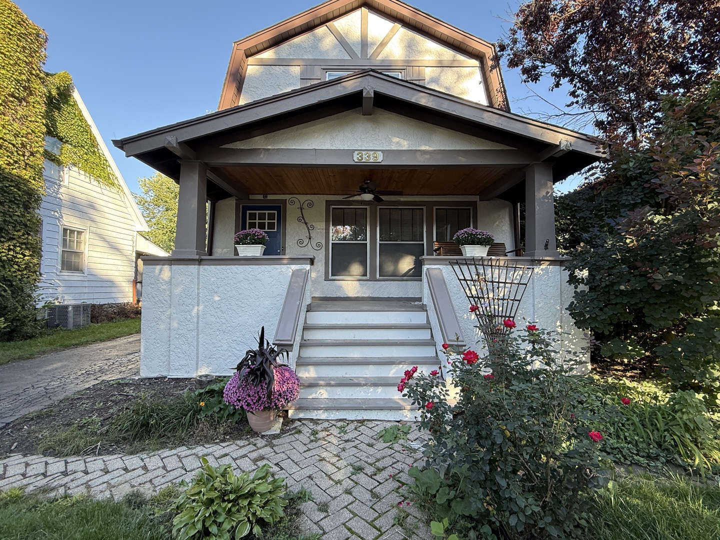 Front exterior of a house with a deep covered porch, broad steps, and flower gardens.