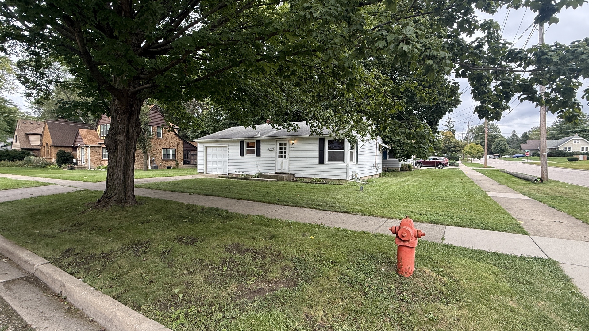Single-story white house with a garage and dark shutters beneath a large leafy tree.
