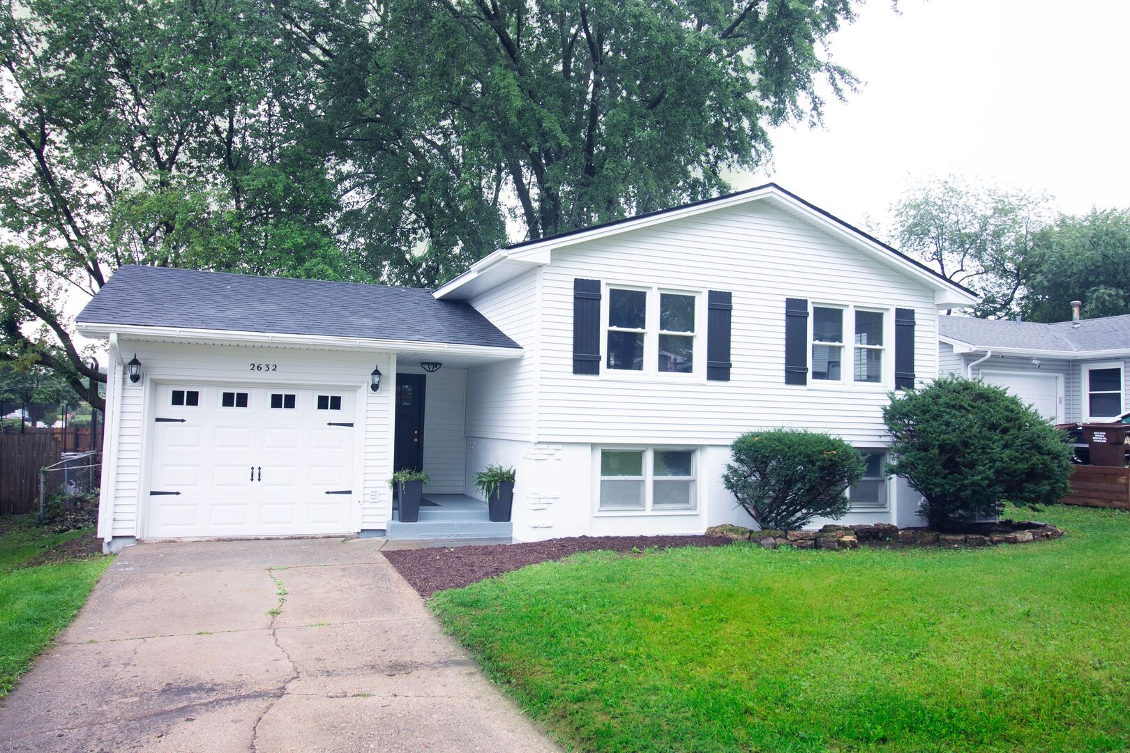White split-level house with black shutters, an attached garage, and mature trees.