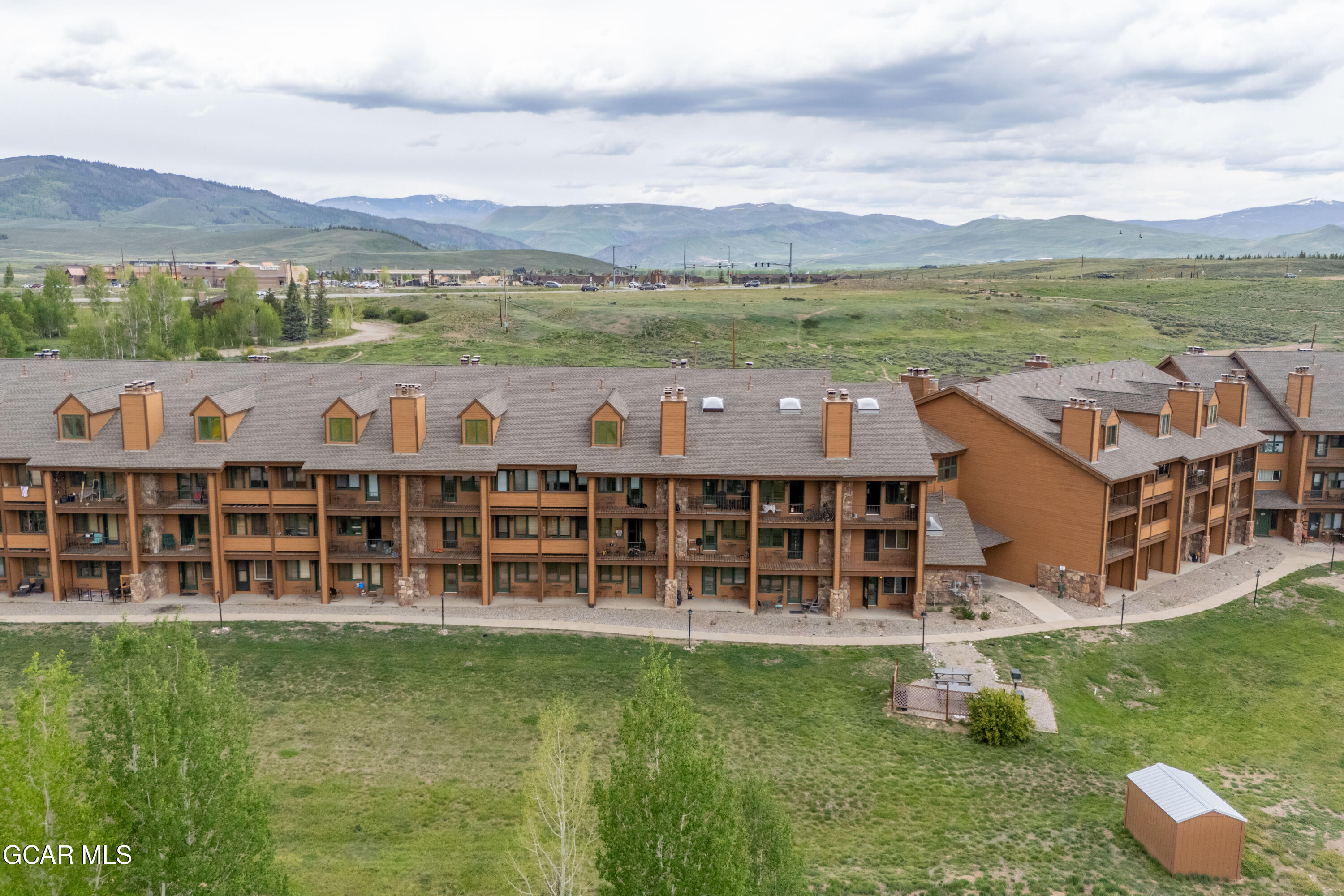 Aerial view of a multi-story condominium building with balconies, grassy grounds, and mountains beyond.