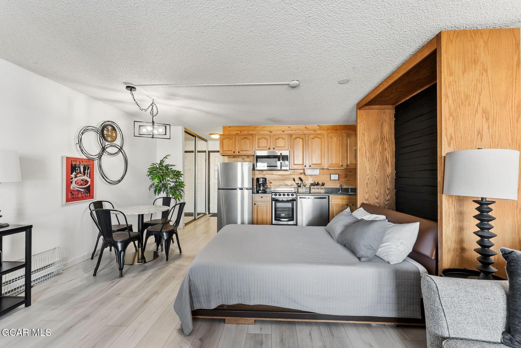 Furnished studio bedroom with a large bed, dining nook, and compact wood-cabinet kitchen.