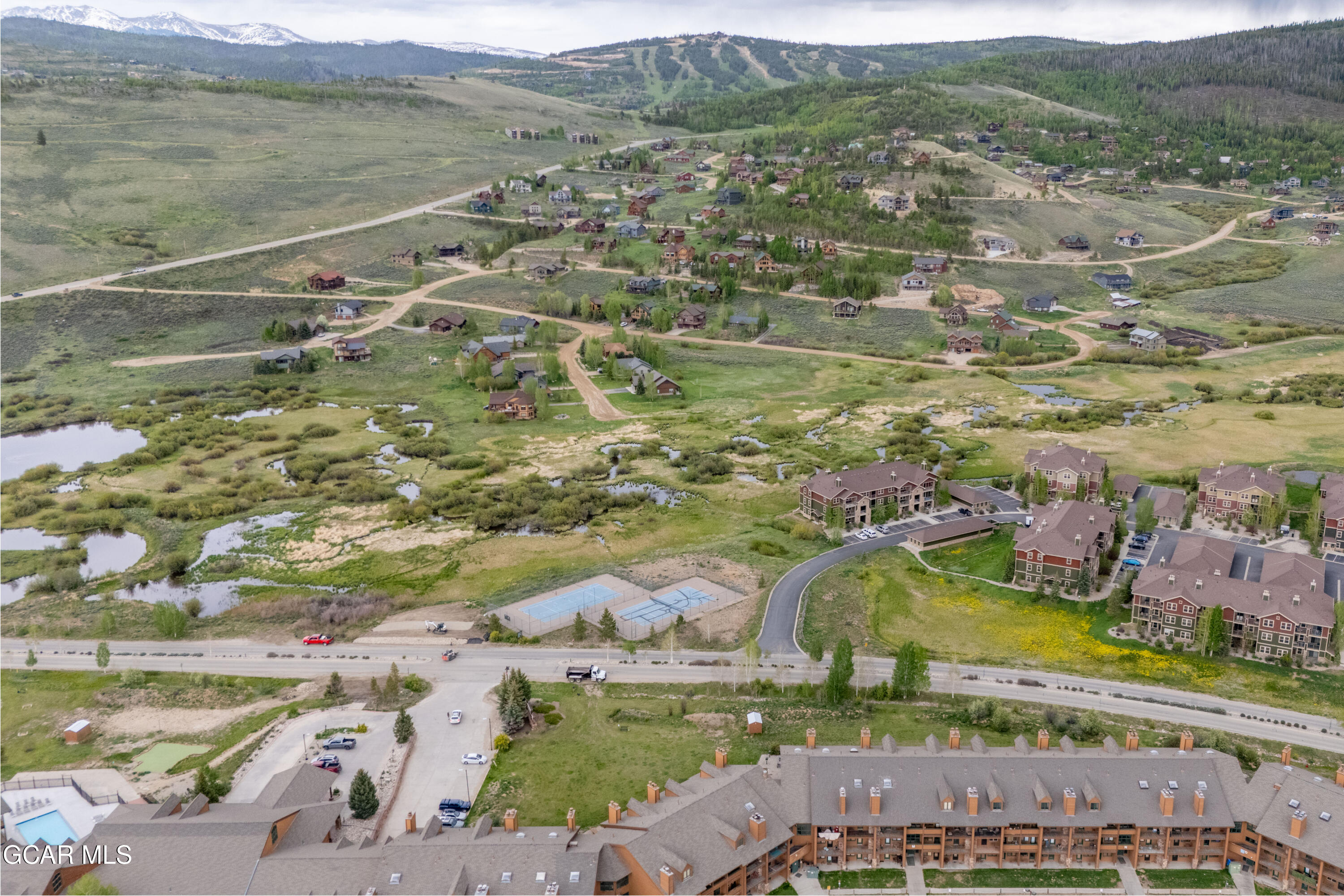 Aerial view of a condominium complex beside wetlands and tennis courts, with mountains in the distance.