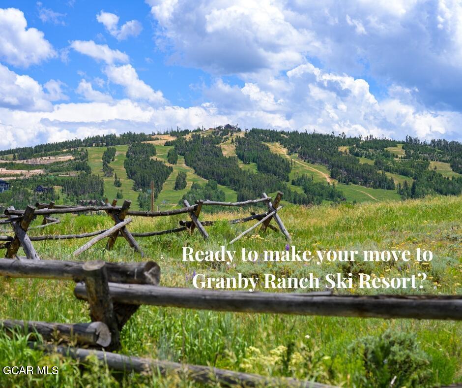 Grassy ski slopes beyond a rustic split-rail fence under a bright, cloud-filled sky.