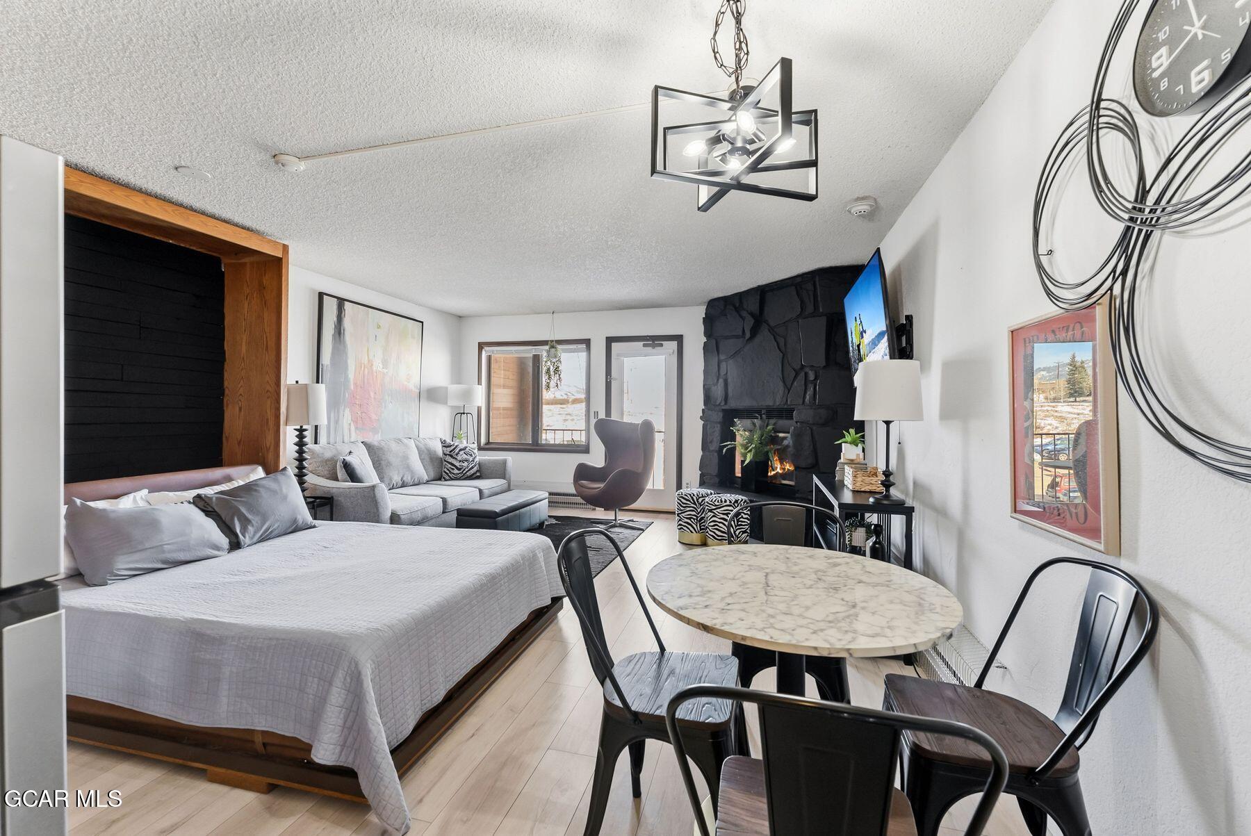 Furnished studio with a bed, dining table, and lit stone fireplace beside a snowy view.