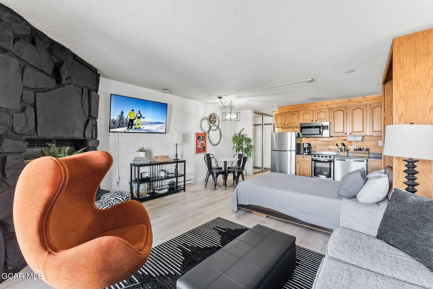 Furnished studio with a black stone fireplace, orange chair, bed, gray sofa, and open kitchen.