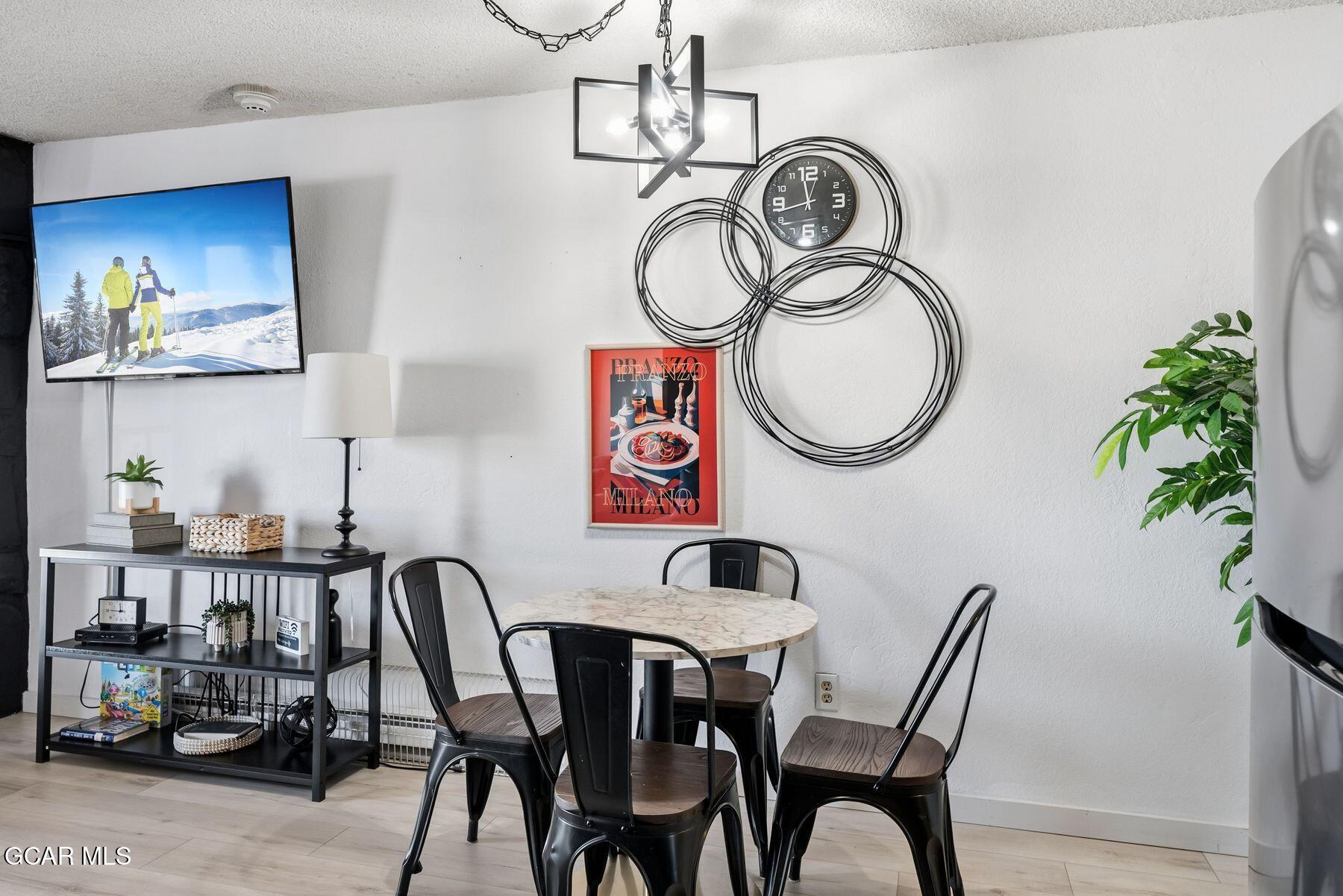 Dining area with a round table, black metal chairs, sculptural wall clock, and television.