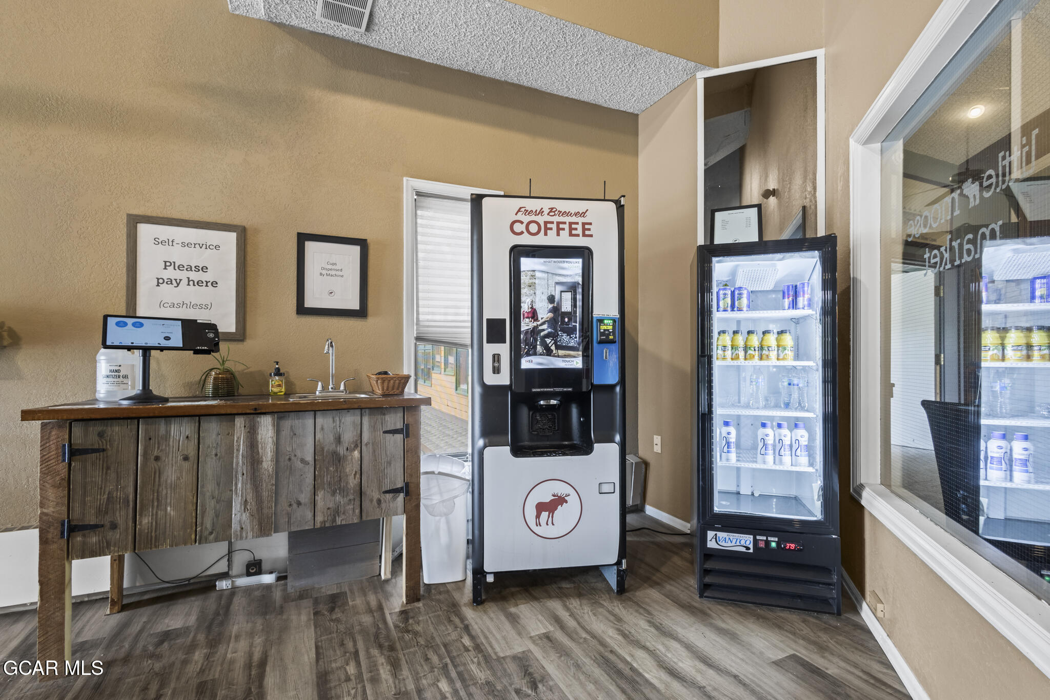 Self-service coffee station with a touch-screen coffee machine, rustic counter, and stocked beverage refrigerator.