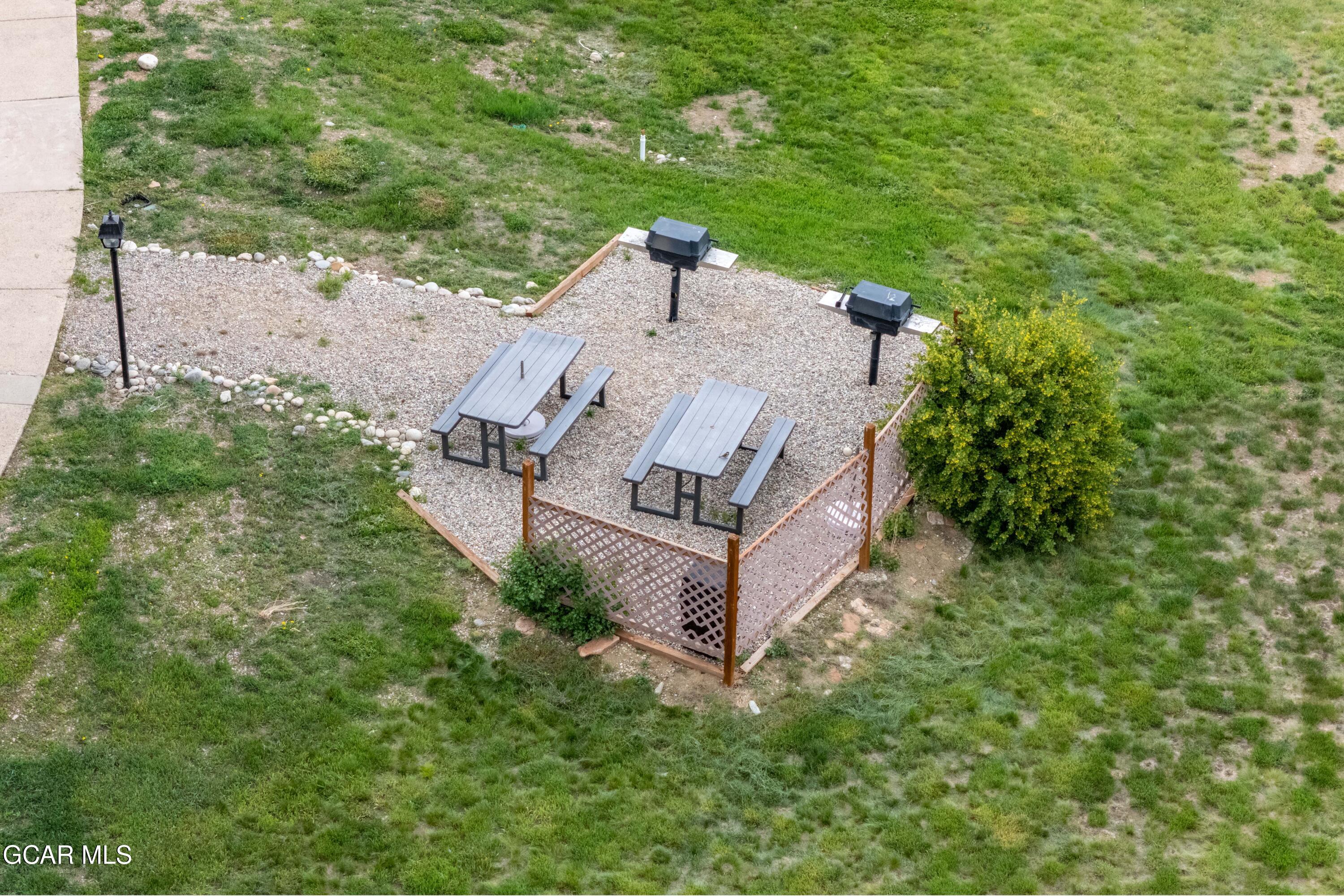 Gravel patio with two picnic tables and grills, bordered by lattice fencing and lawn.