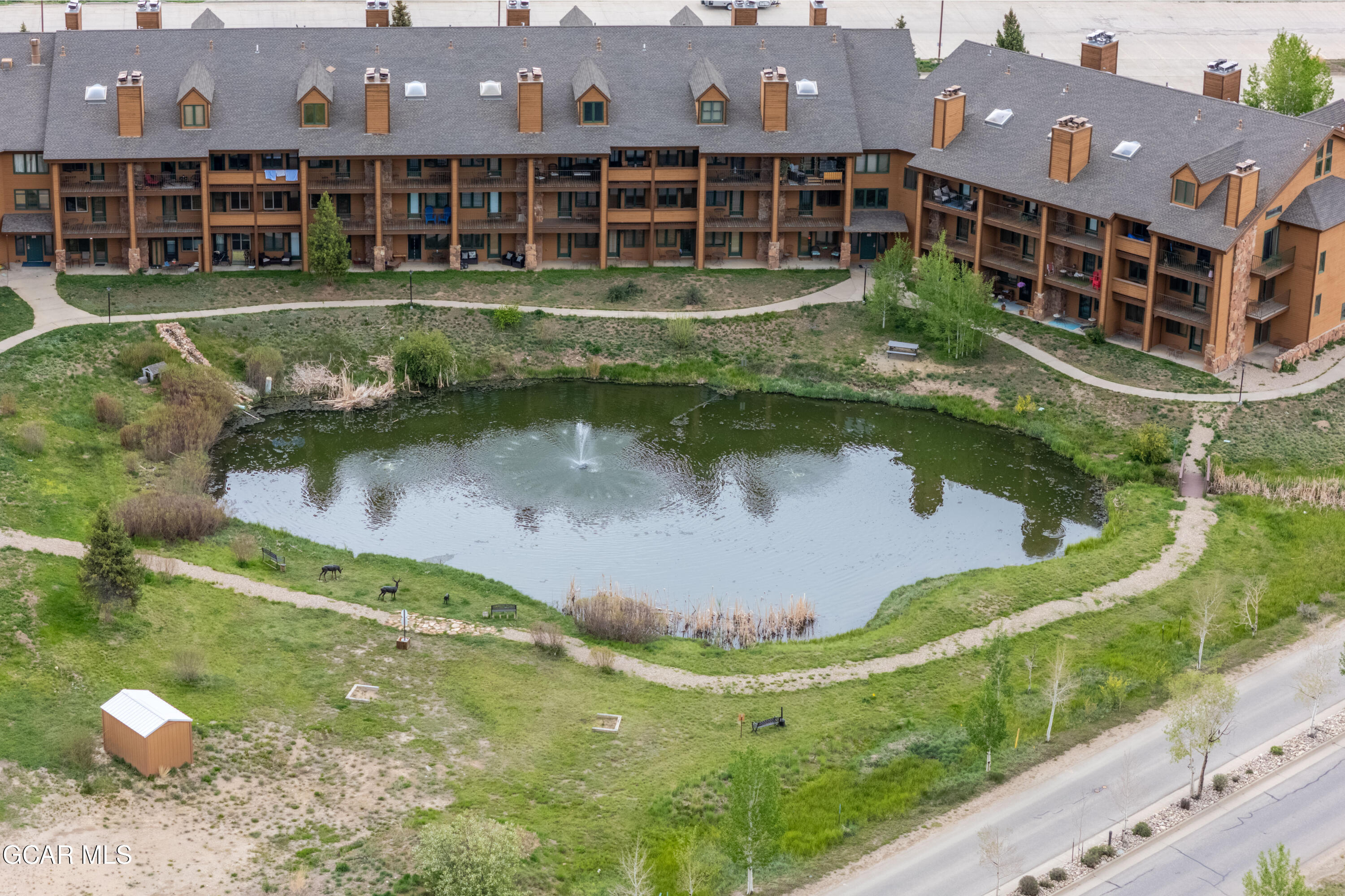 Aerial view of a brown condominium building beside a pond with a fountain and two elk.