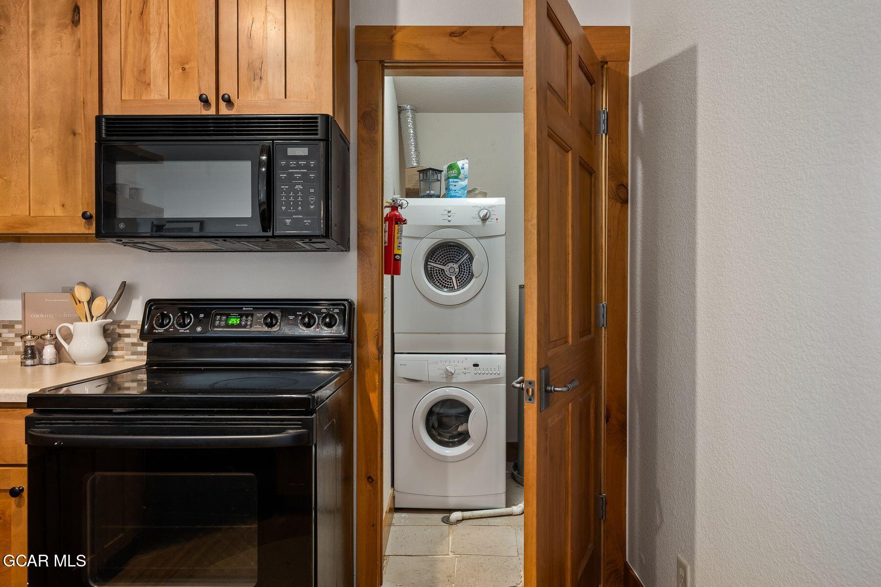 Kitchen with wood cabinets and black appliances beside an open laundry closet with a stacked washer and dryer.