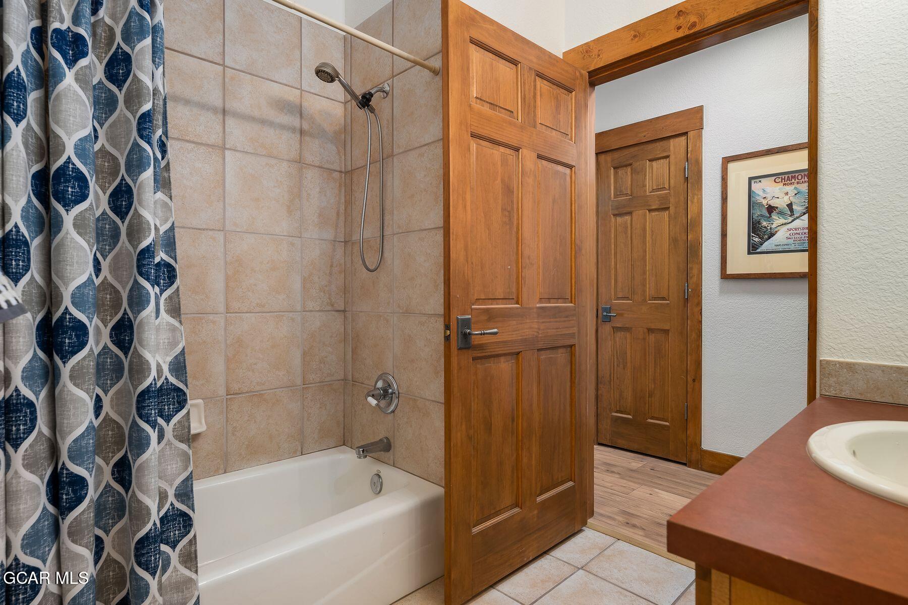 Bathroom with a tiled tub and shower, patterned curtain, wood doors, and vanity.