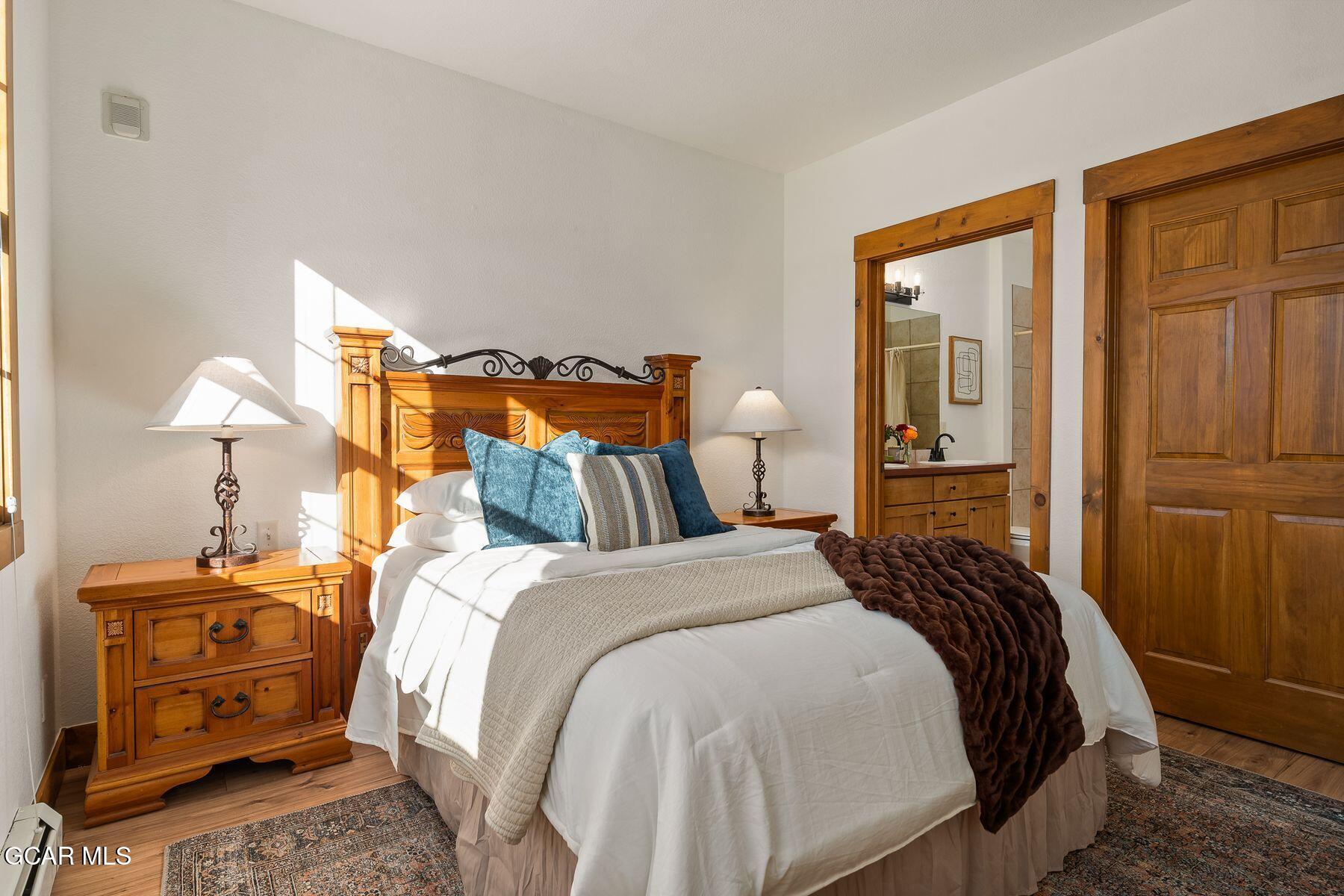 Sunlit bedroom with white bedding, blue pillows, ornate wood furniture, and an ensuite bathroom doorway.