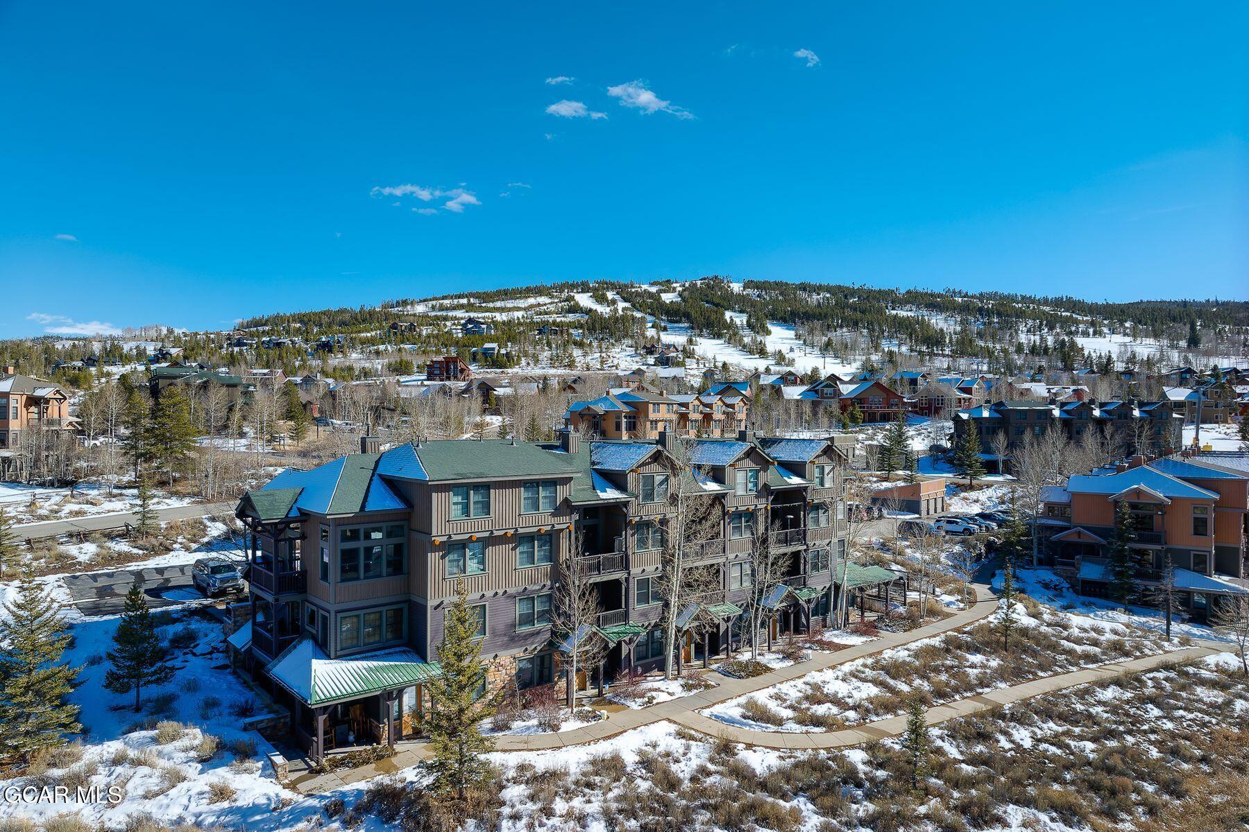 Condominium buildings below a snowy, tree-lined ski slope under a clear blue sky.
