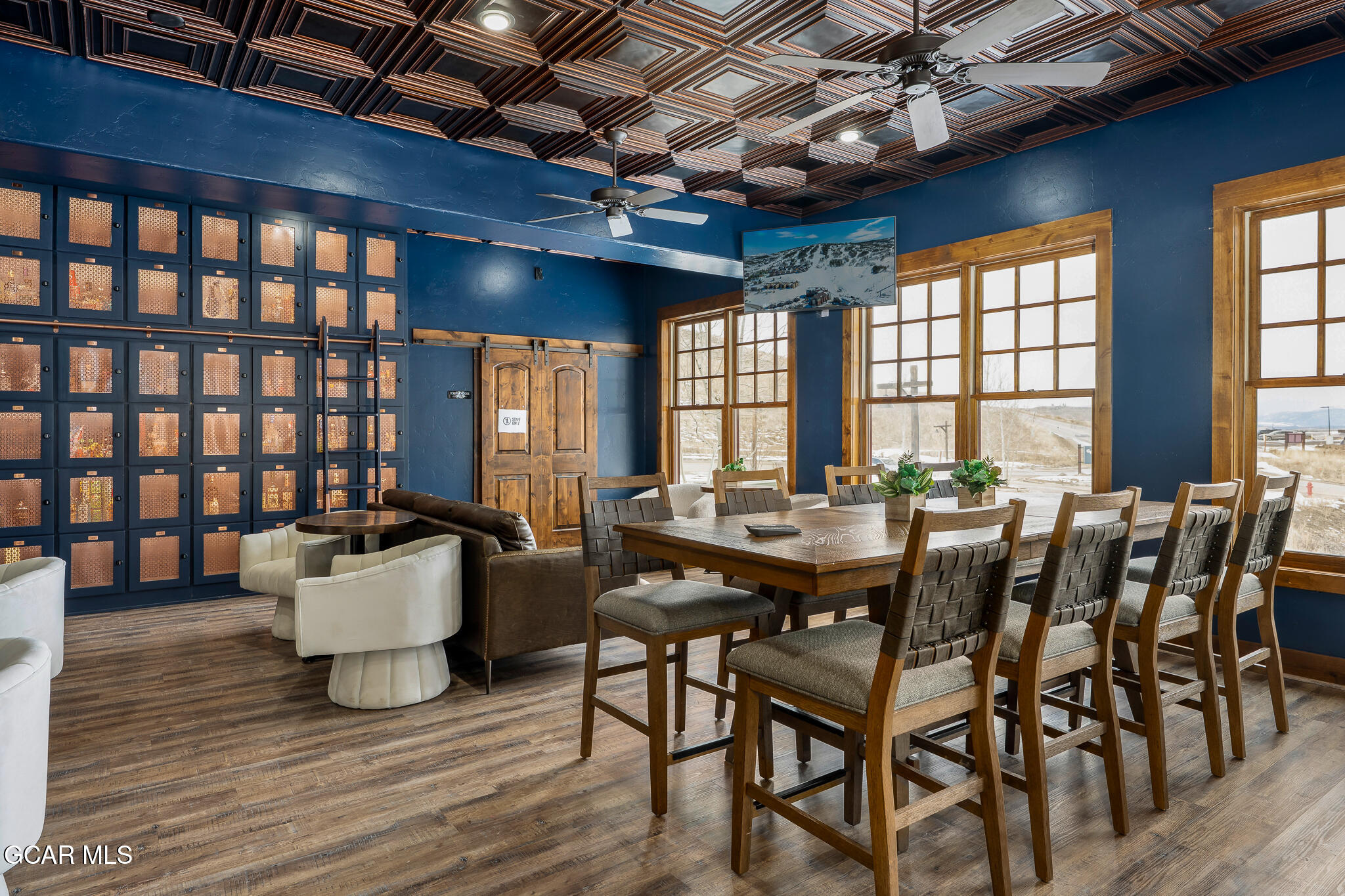 Communal dining lounge with a long wooden table, blue walls, coffered ceiling, and windows overlooking snow.