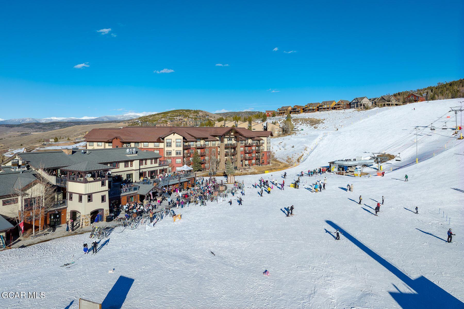 Snowy ski slope with skiers, lift equipment, resort buildings, and distant mountains.