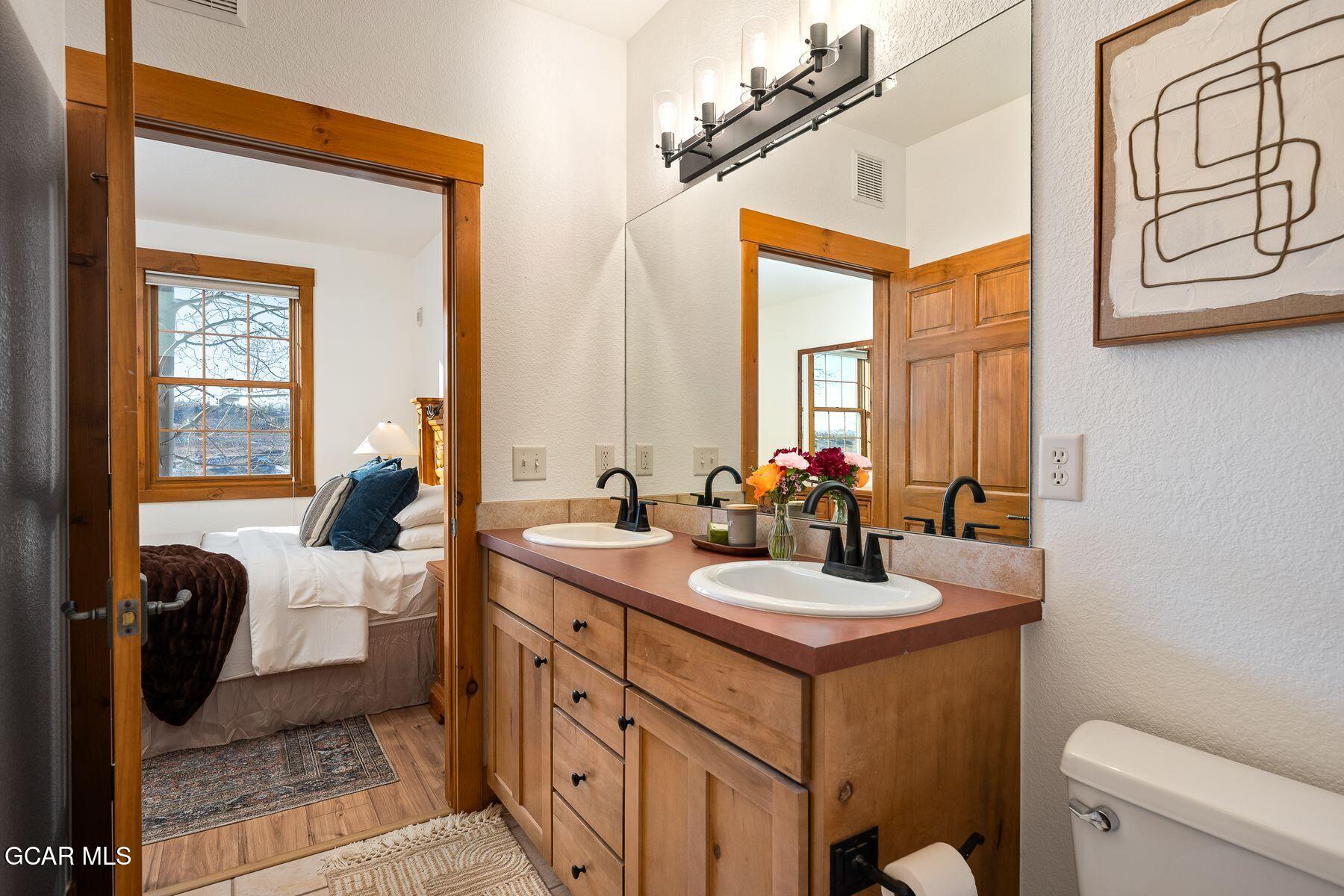 Bathroom with a double-sink wood vanity and a view through the doorway into a furnished bedroom.
