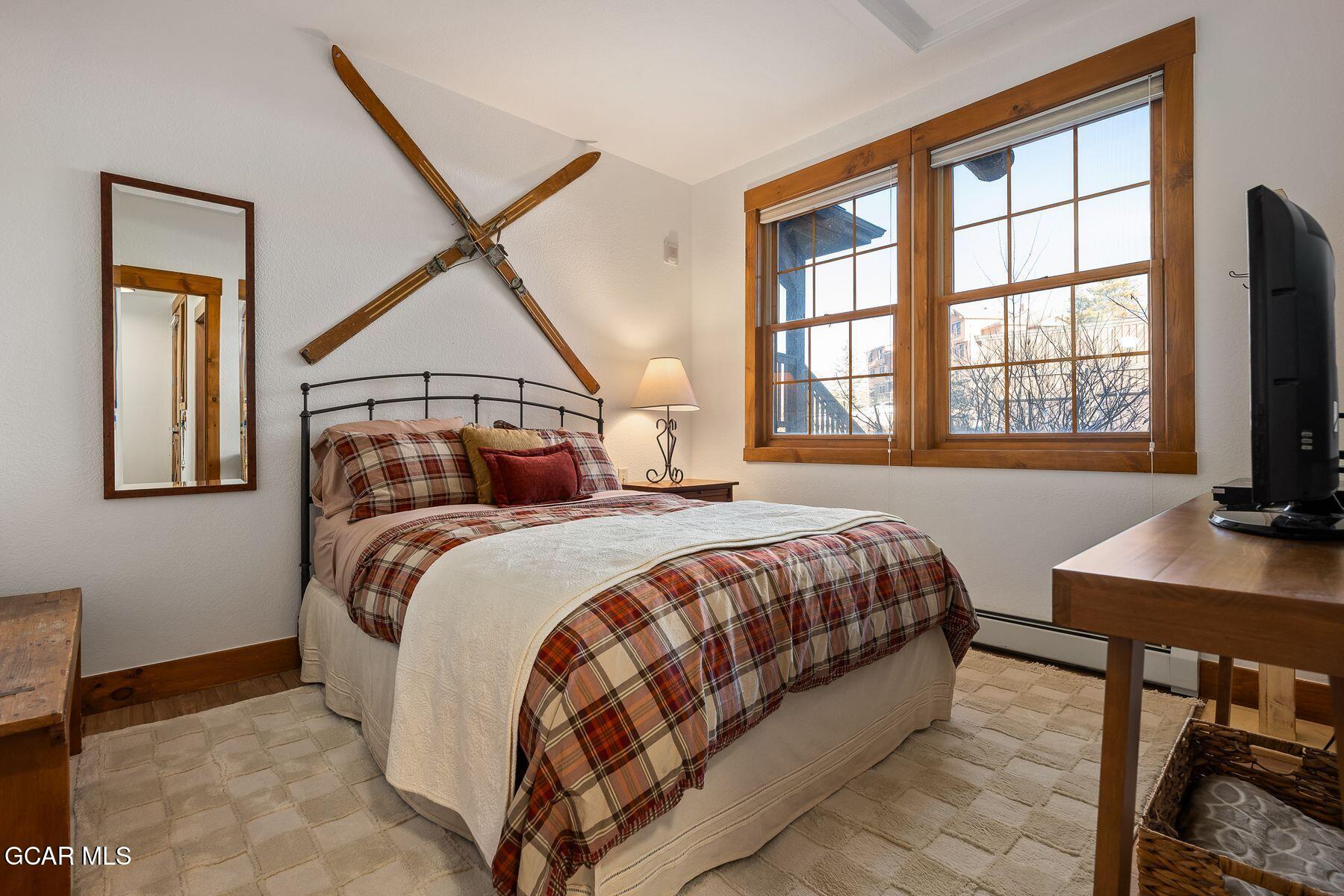 Bedroom with plaid bedding, crossed vintage skis above the bed, and large wood-trimmed windows.