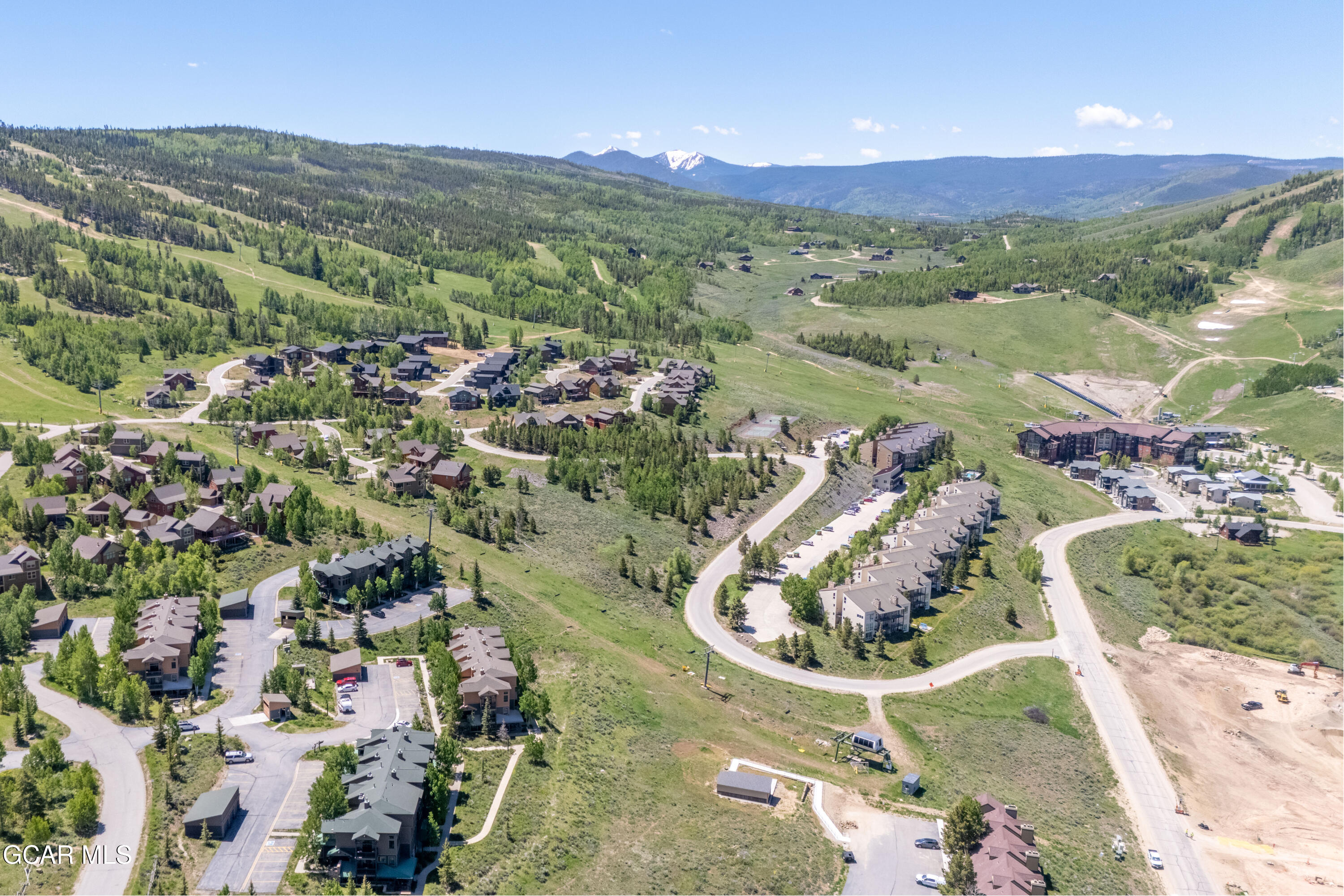 Aerial view of a mountain residential development with winding roads, forested hills, and distant snowy peaks.