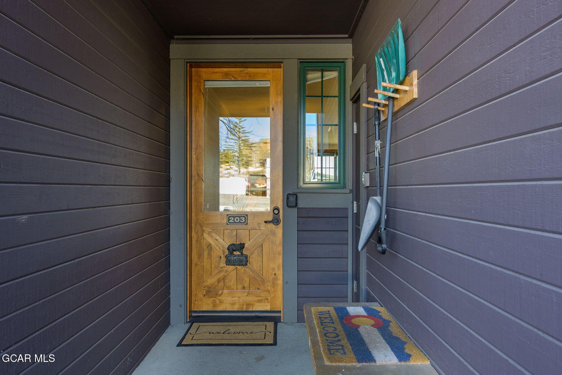 Covered entry porch with a wood-and-glass door marked 203, Colorado flag, and welcome mats.